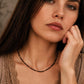 Close-up lifestyle view of a model wearing a dainty black spinel choker necklace with small 22k gold vermeil accent beads. The minimalist beaded necklace rests perfectly on her collarbone, styled alongside a neutral beige top with delicate lace detailing. The shot showcases the elegant scale and fit of the gemstone jewellery on the wrist and neck.