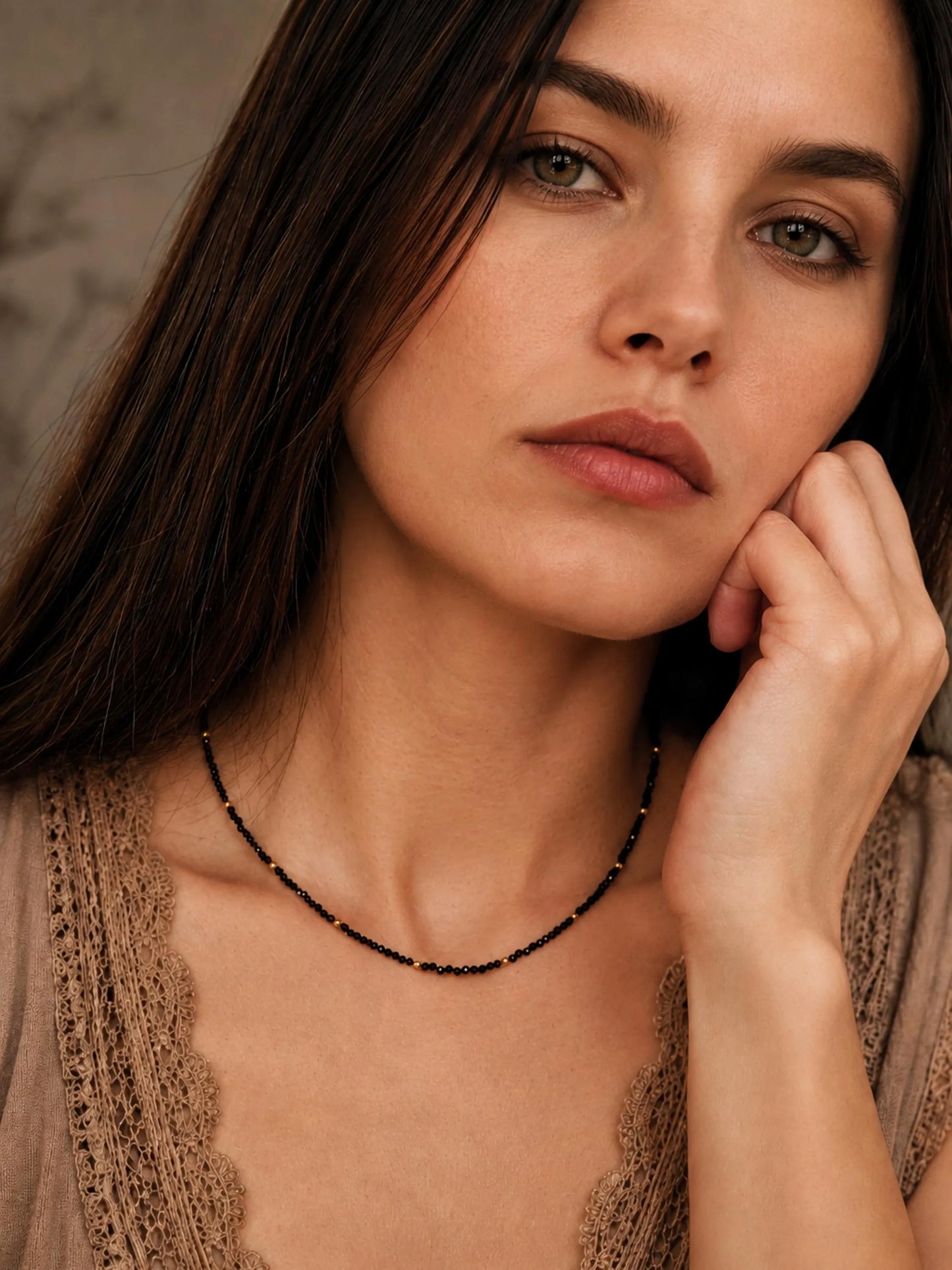 Close-up lifestyle view of a model wearing a dainty black spinel choker necklace with small 22k gold vermeil accent beads. The minimalist beaded necklace rests perfectly on her collarbone, styled alongside a neutral beige top with delicate lace detailing. The shot showcases the elegant scale and fit of the gemstone jewellery on the wrist and neck.
