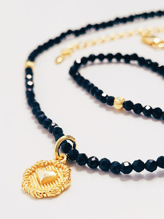 Close-up of a black spinel beaded choker with a gold puff heart pendant, featuring extra shine black beads and intricate gold detailing. The necklace lies on a white background, showcasing its elegance and refined craftsmanship.