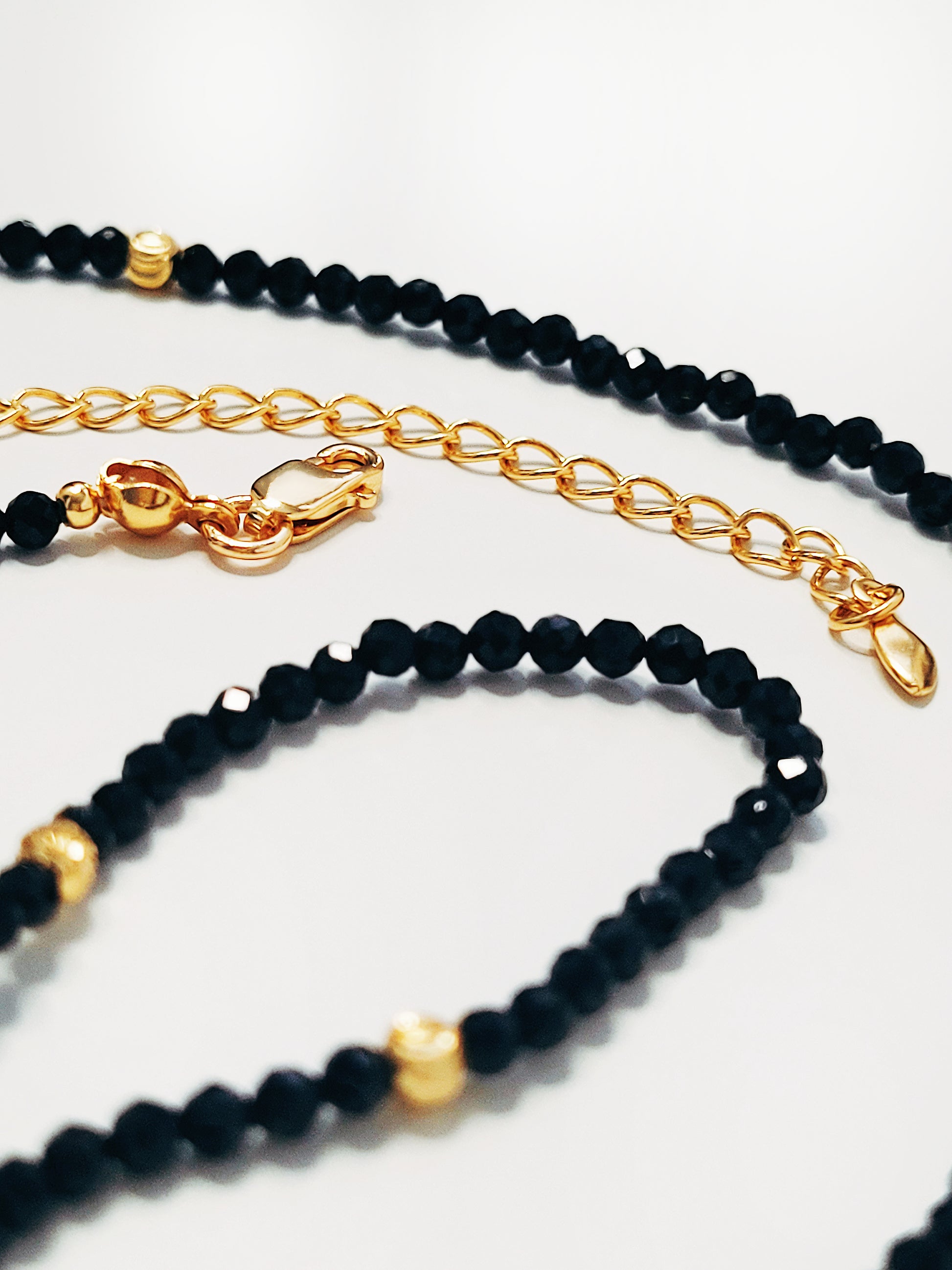 Close-up of the lobstee clasp closure of a black spinel beaded choker with a gold puff heart pendant, featuring extra shine black beads and intricate gold detailing lying on a white background, showcasing its elegance and refined craftsmanship.