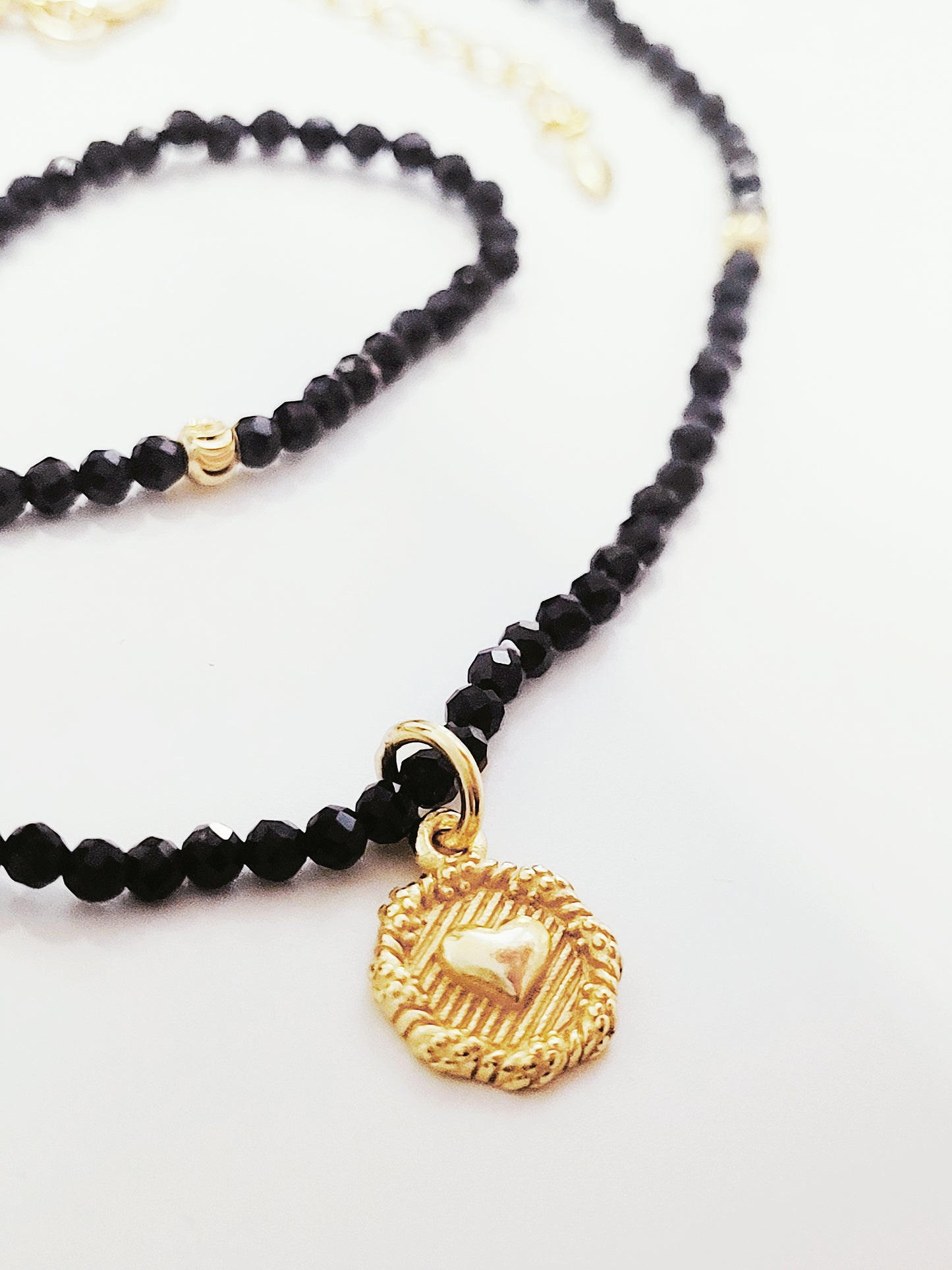 Close-up of a black spinel beaded choker with a gold puff heart pendant, featuring extra shine black beads and intricate gold detailing. The necklace lies on a white background, showcasing its elegance and refined craftsmanship.