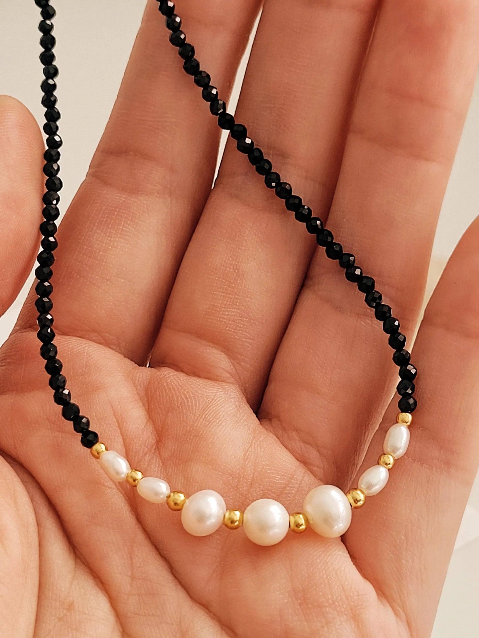 Close-up of a hand holding a black spinel beaded gemstone choker necklace with freshwater pearls again an aesthetic luxury beige background.