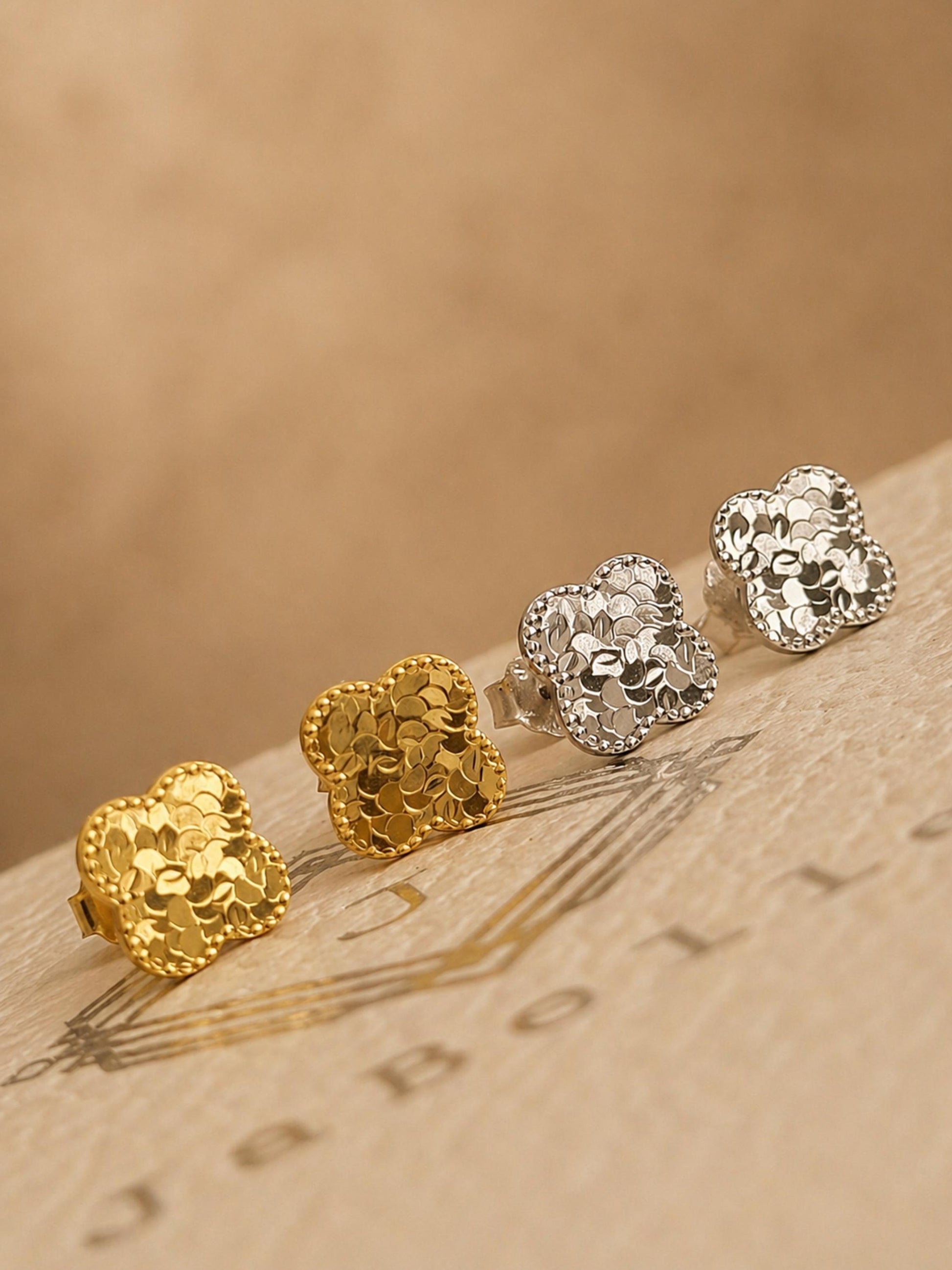 Close up of a set of dainty clover stud earrings in 18k gold vermeil and 925 sterling silver showing the intricate textured detail on an aesthetic beige stone surface, perfect gift for her