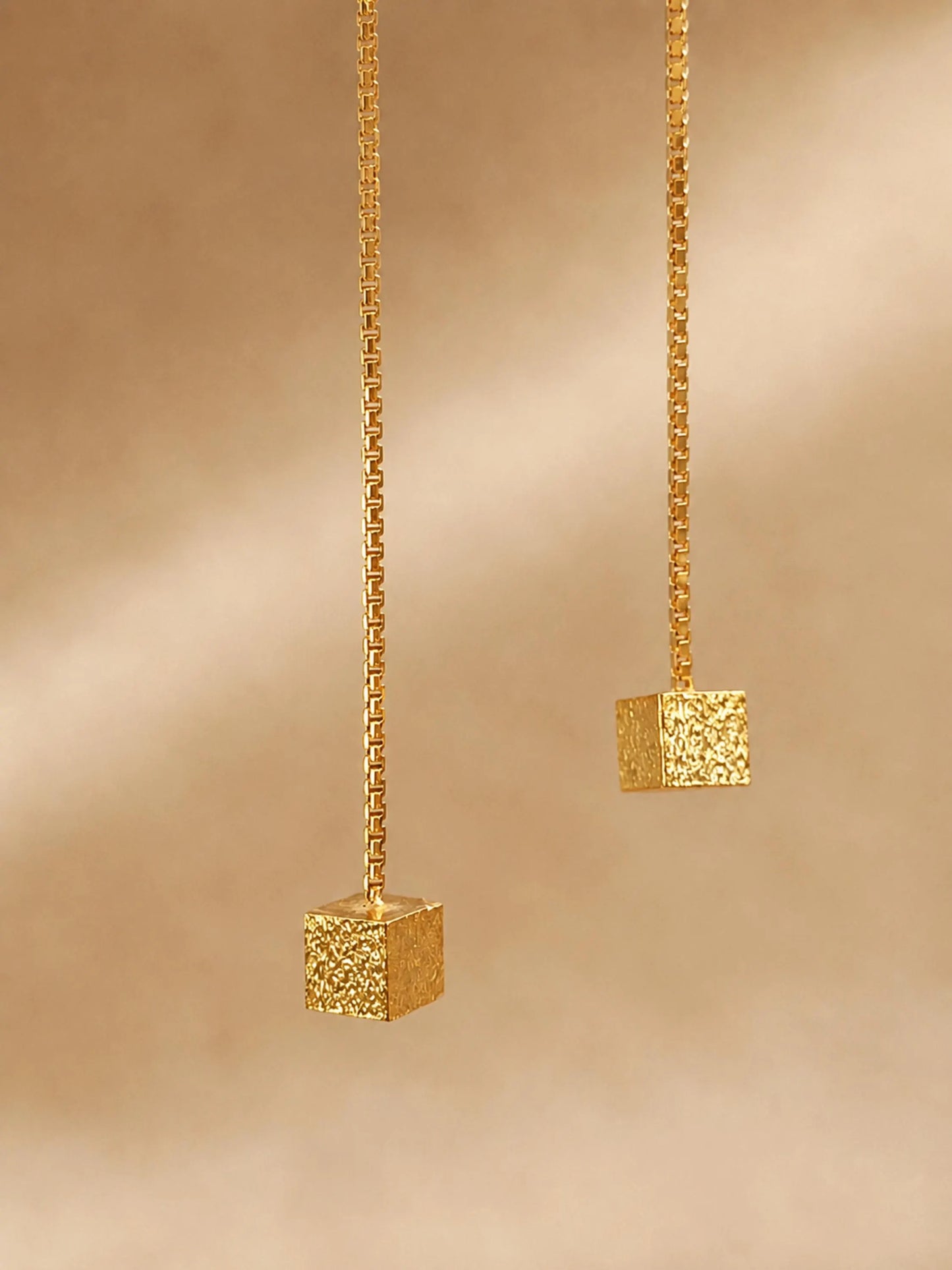 Close-up of two gold box chain threader earrings showing the fine detail of the textured, glittering square cube charms suspended at the ends against a warm, neutral background.