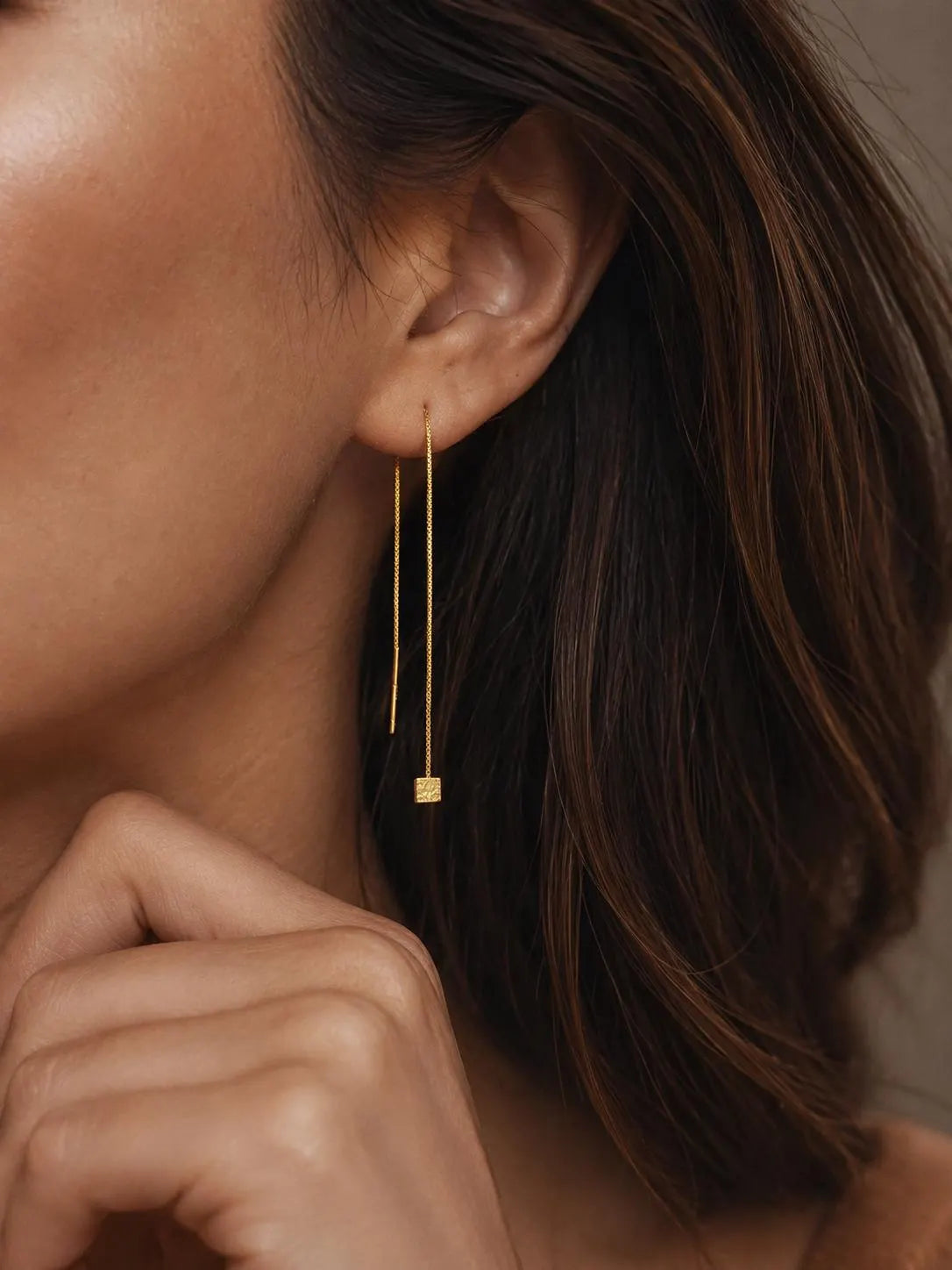 A woman with dark hair wearing a single gold box chain threader earring with a small geometric cube charm hanging elegantly below her earlobe.