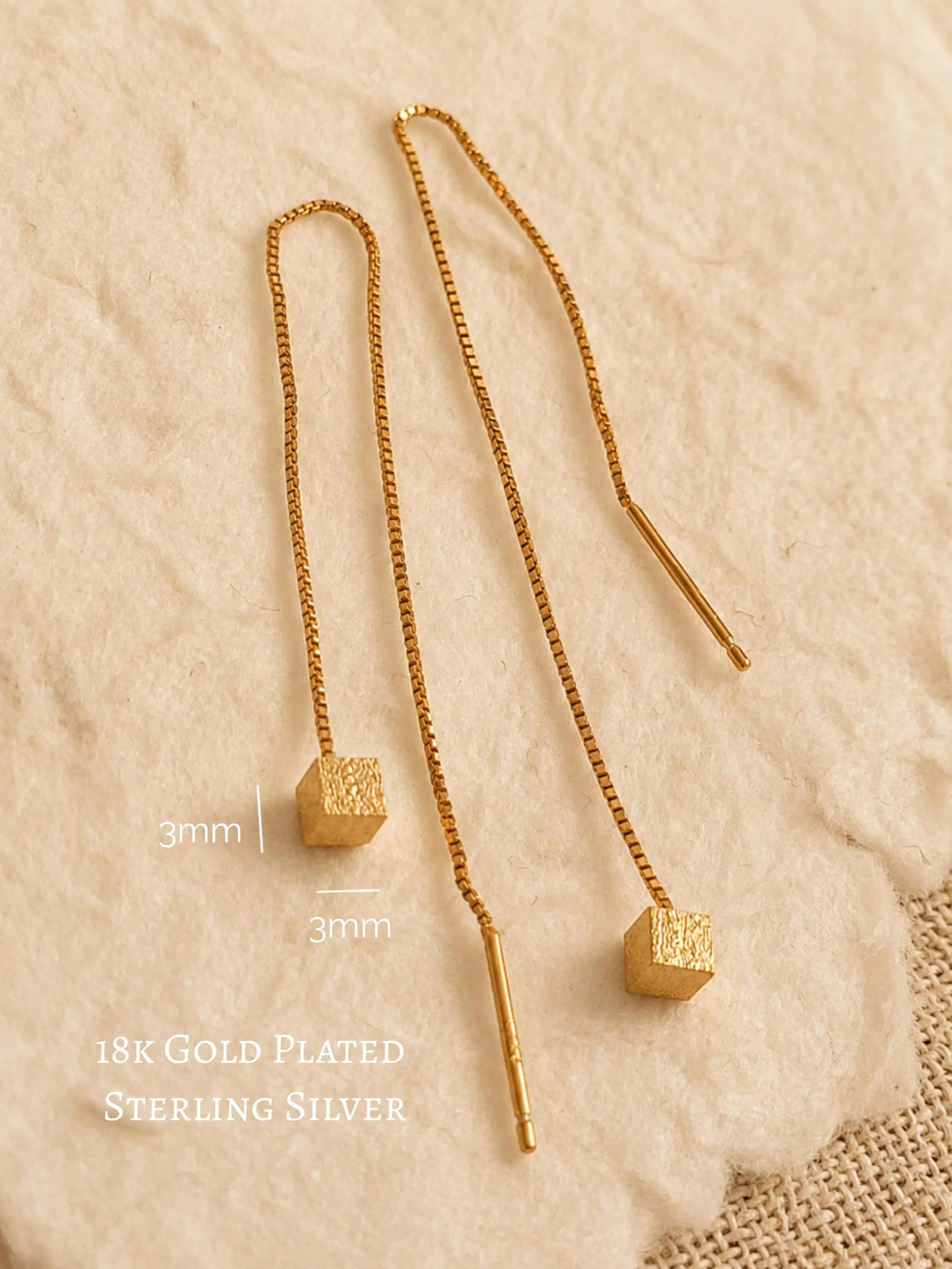 Close-up of two gold box chain threader earrings showing the fine detail of the textured, glittering square cube charms suspended at the ends against a warm, neutral background.