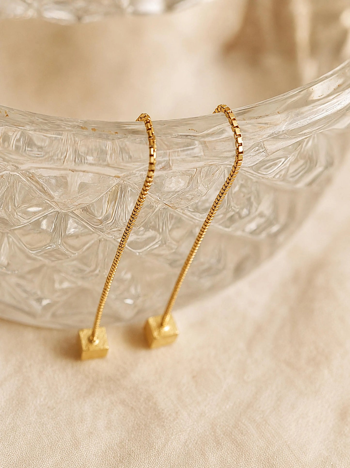 Close-up of two gold box chain threader earrings showing the fine detail of the textured, glittering square cube charms suspended at the ends against a warm, neutral background.