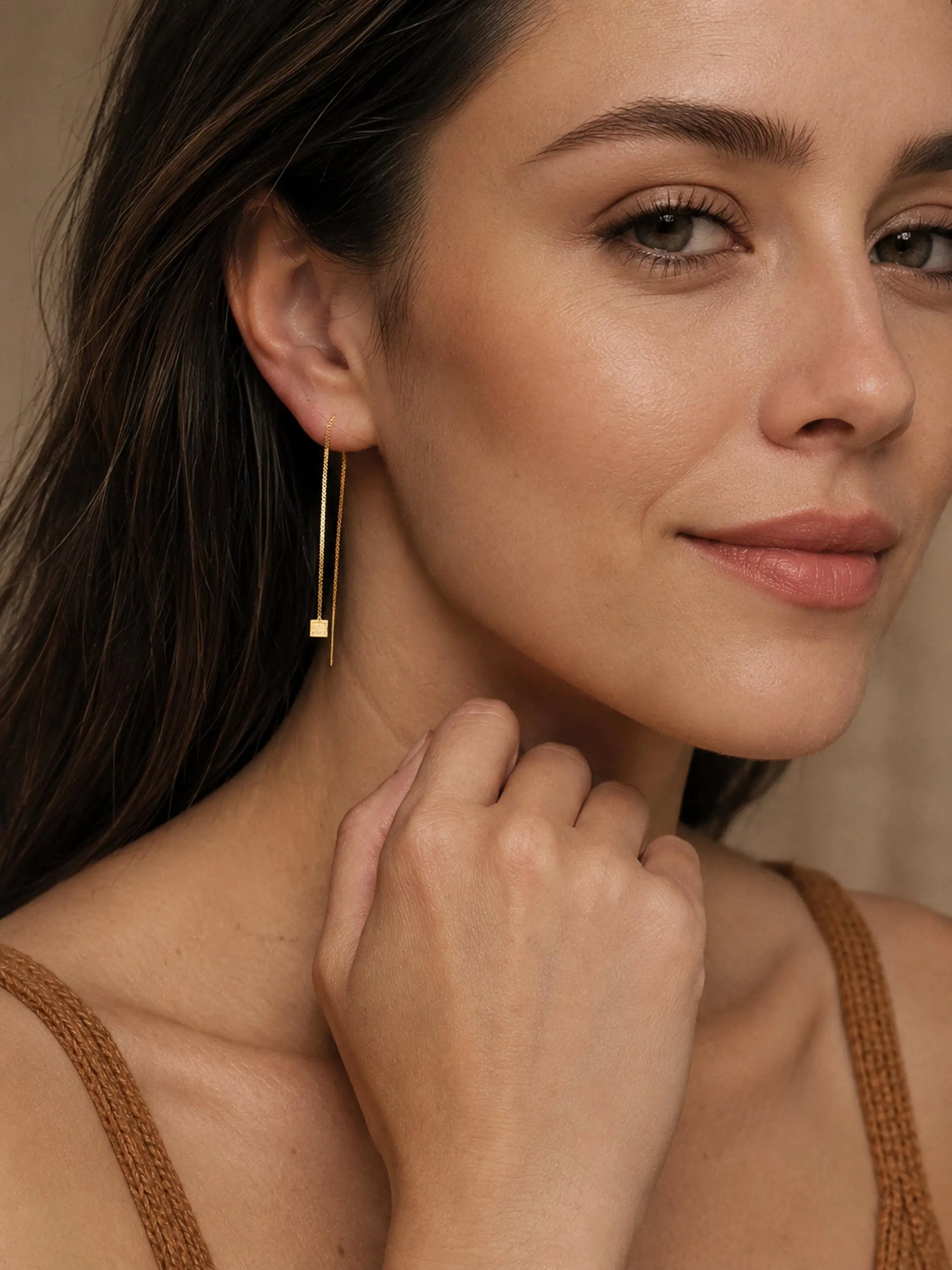 A woman with curl brown hair wearing a gold box chain threader earring pull through with a small geometric cube charm hanging elegantly below her earlobe.