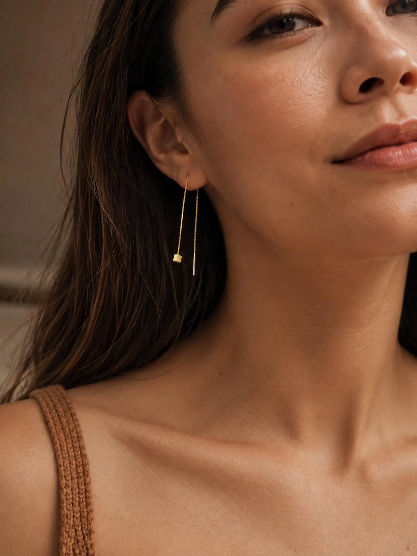 A woman with dark hair wearing a single gold box chain threader earring with a small geometric cube charm hanging elegantly below her earlobe.