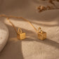 Close-up of two gold box chain threader earrings showing the fine detail of the textured, glittering square cube charms suspended at the ends against a warm, neutral background.