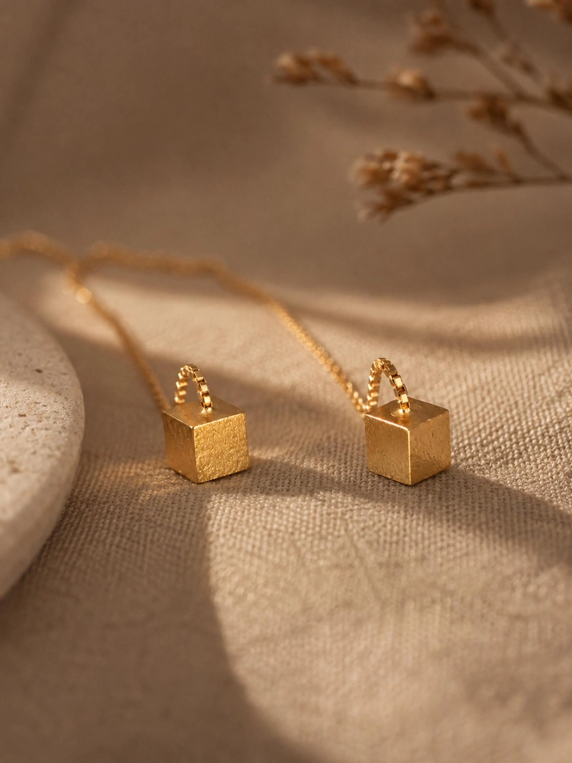 Close-up of two gold box chain threader earrings showing the fine detail of the textured, glittering square cube charms suspended at the ends against a warm, neutral background.
