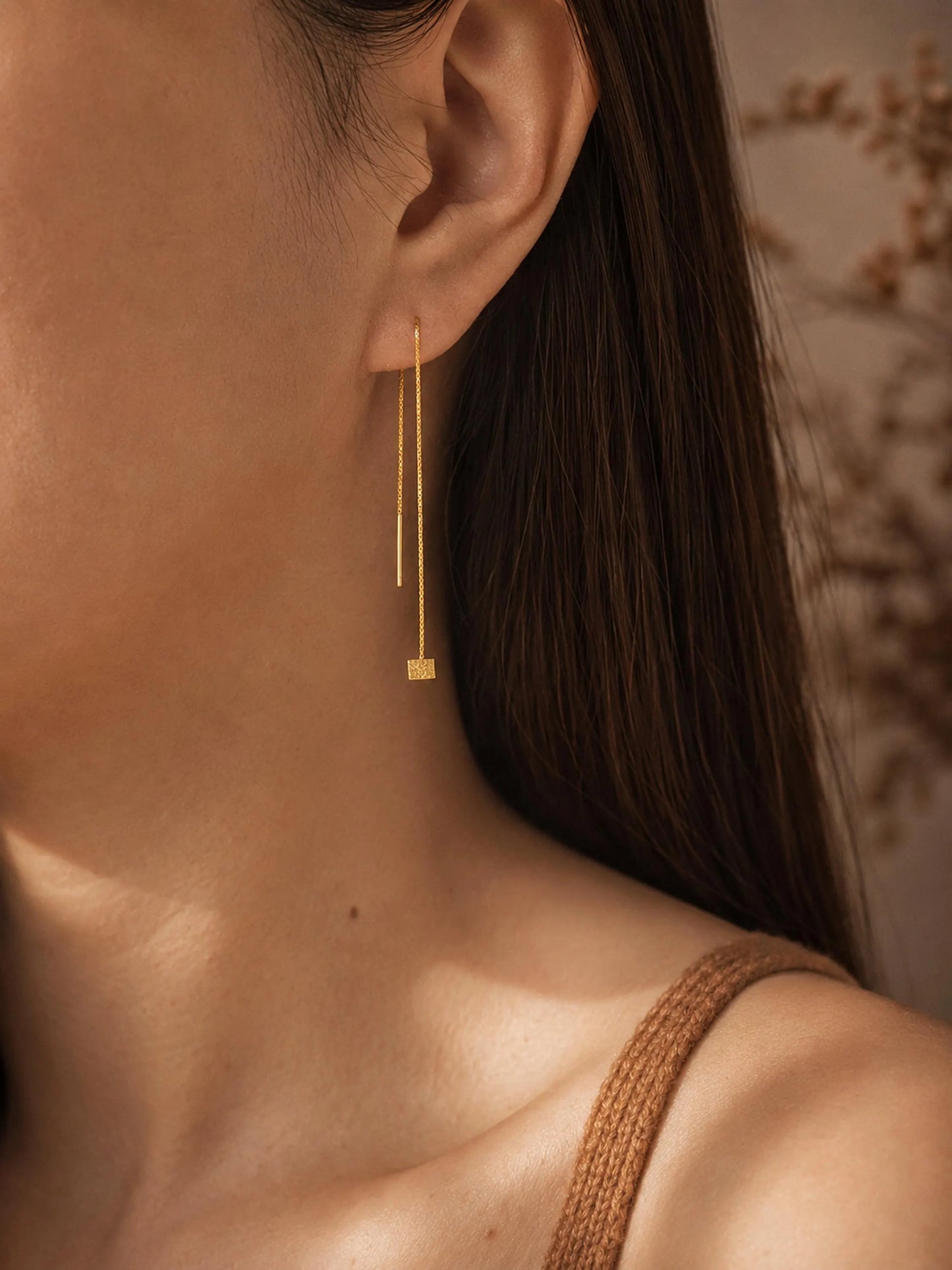 A woman with dark hair wearing a single gold box chain threader earring with a small geometric cube charm hanging elegantly below her earlobe.