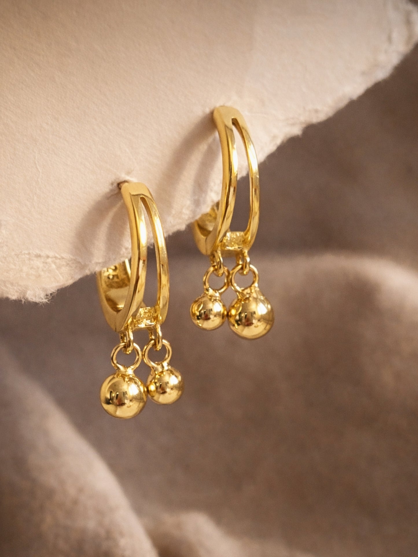 Dainty gold double hoop earrings with dangling balls on a soft beige background, everyday minimalist jewellery.