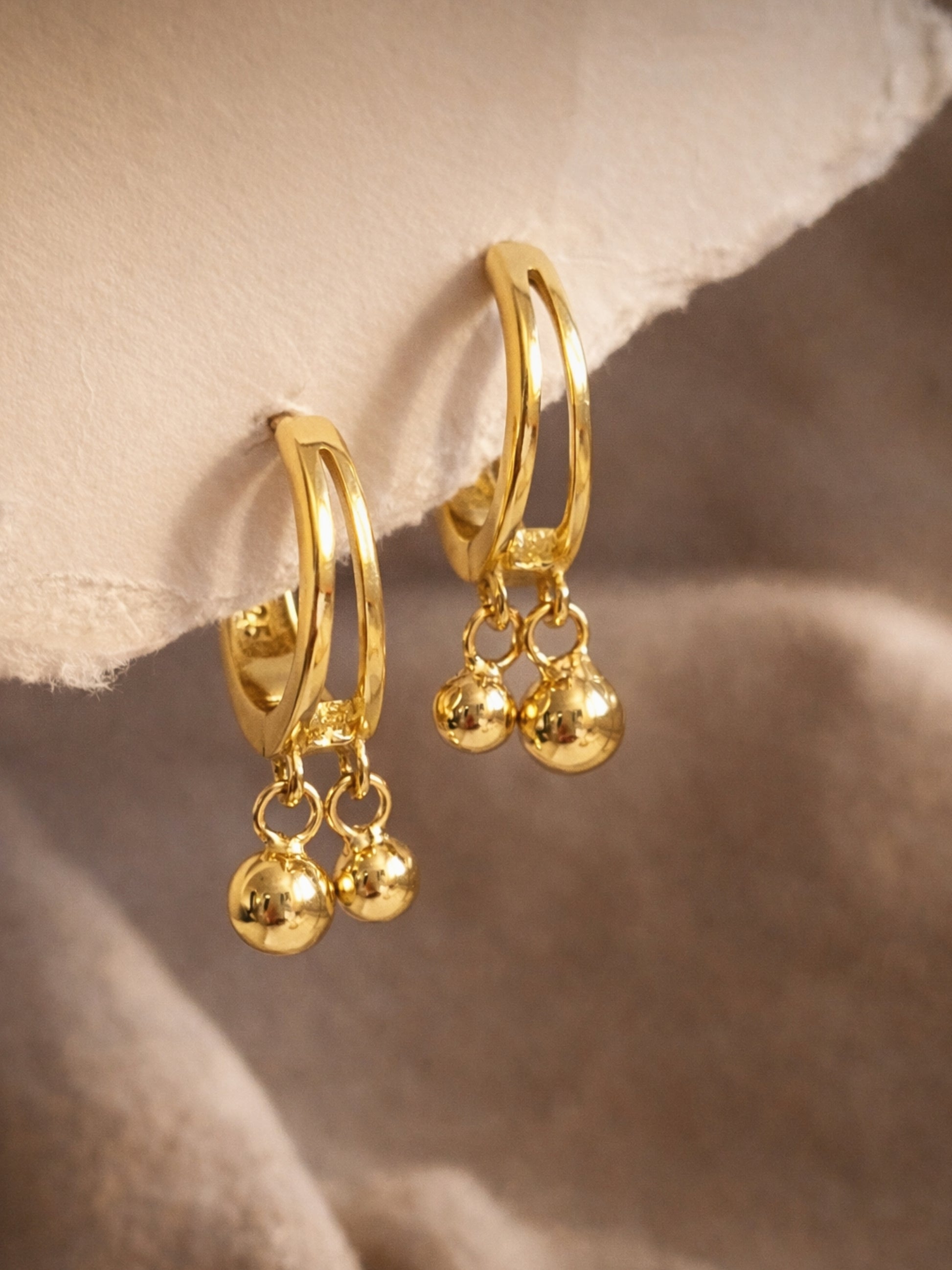 Dainty gold double hoop earrings with dangling balls on a soft beige background, everyday minimalist jewellery.