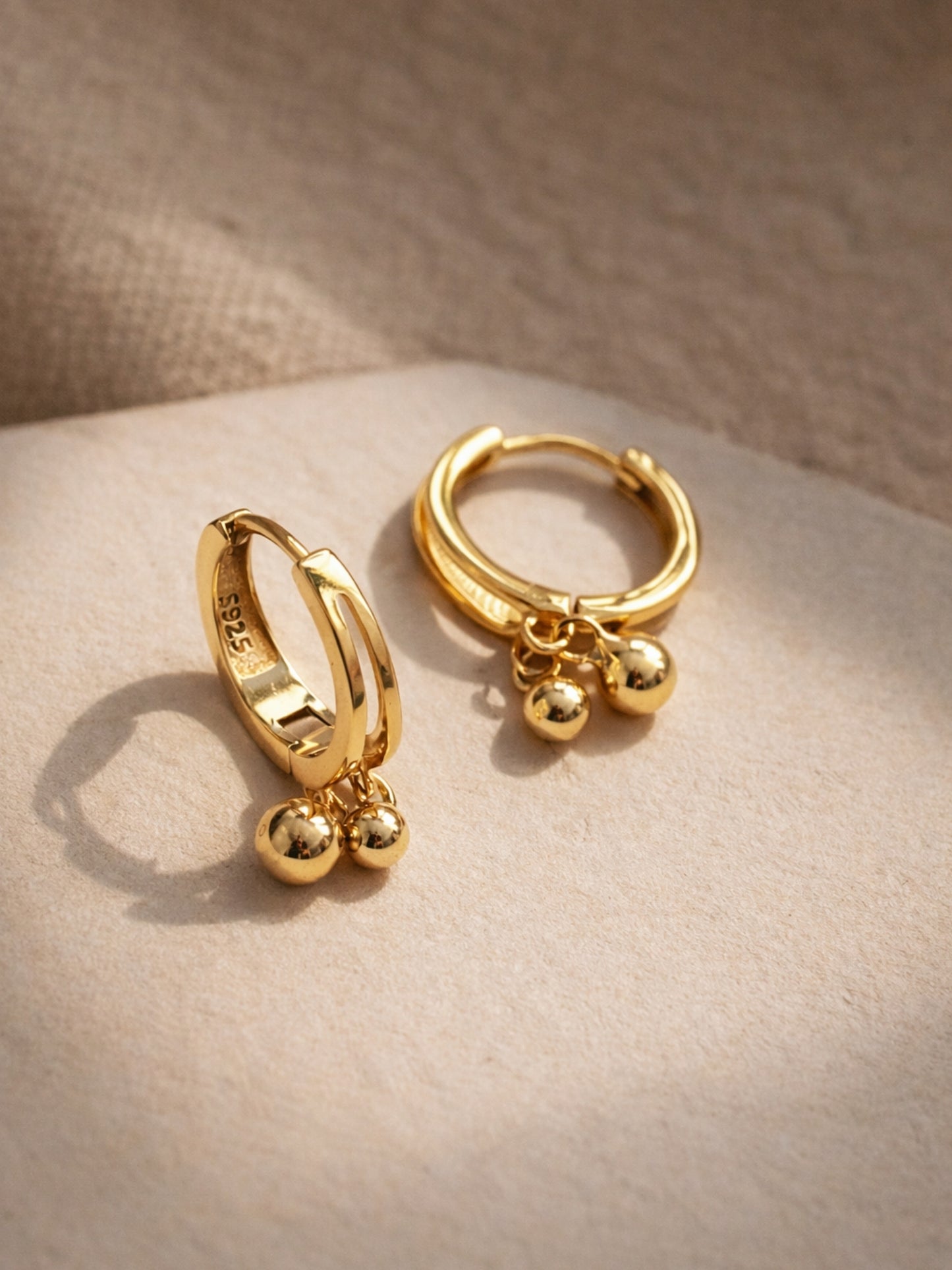 Close up of a pair of gold hoop earrings with spherical charms on a beige surface, crafted in gold plated sterling silver, waterproof and hypoallergenic. 