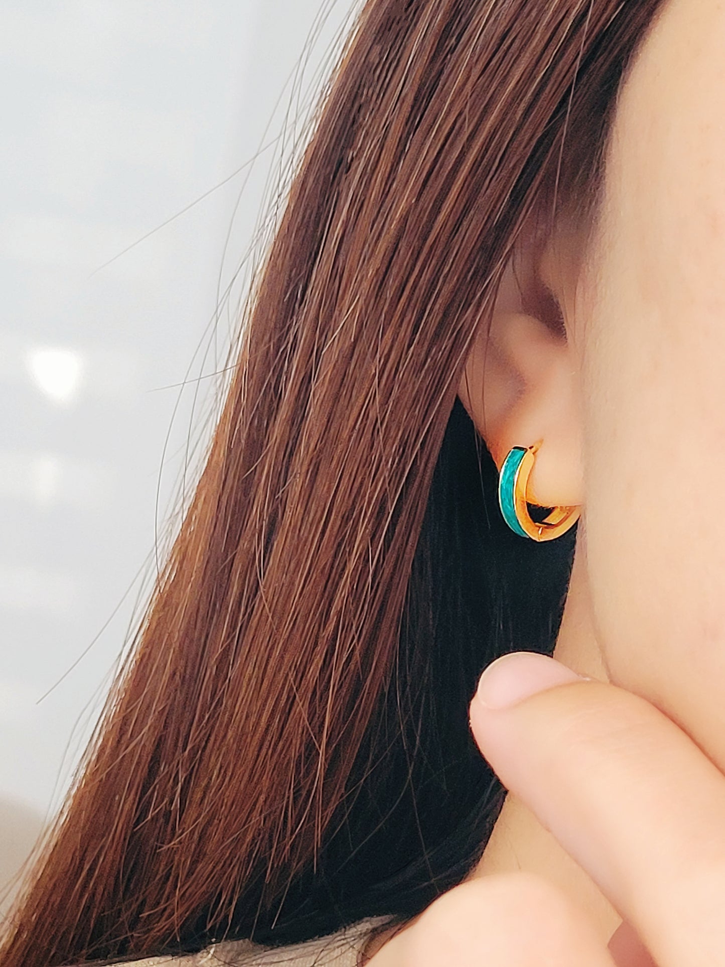 A woman wears eye-catching emeral green enamel huggie hoop earrings, featuring two vibrant colors - turquoise and gold, enhancing her fashionable appearance.