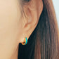 A woman wears eye-catching emeral green enamel huggie hoop earrings, featuring two vibrant colors - turquoise and gold, enhancing her fashionable appearance.