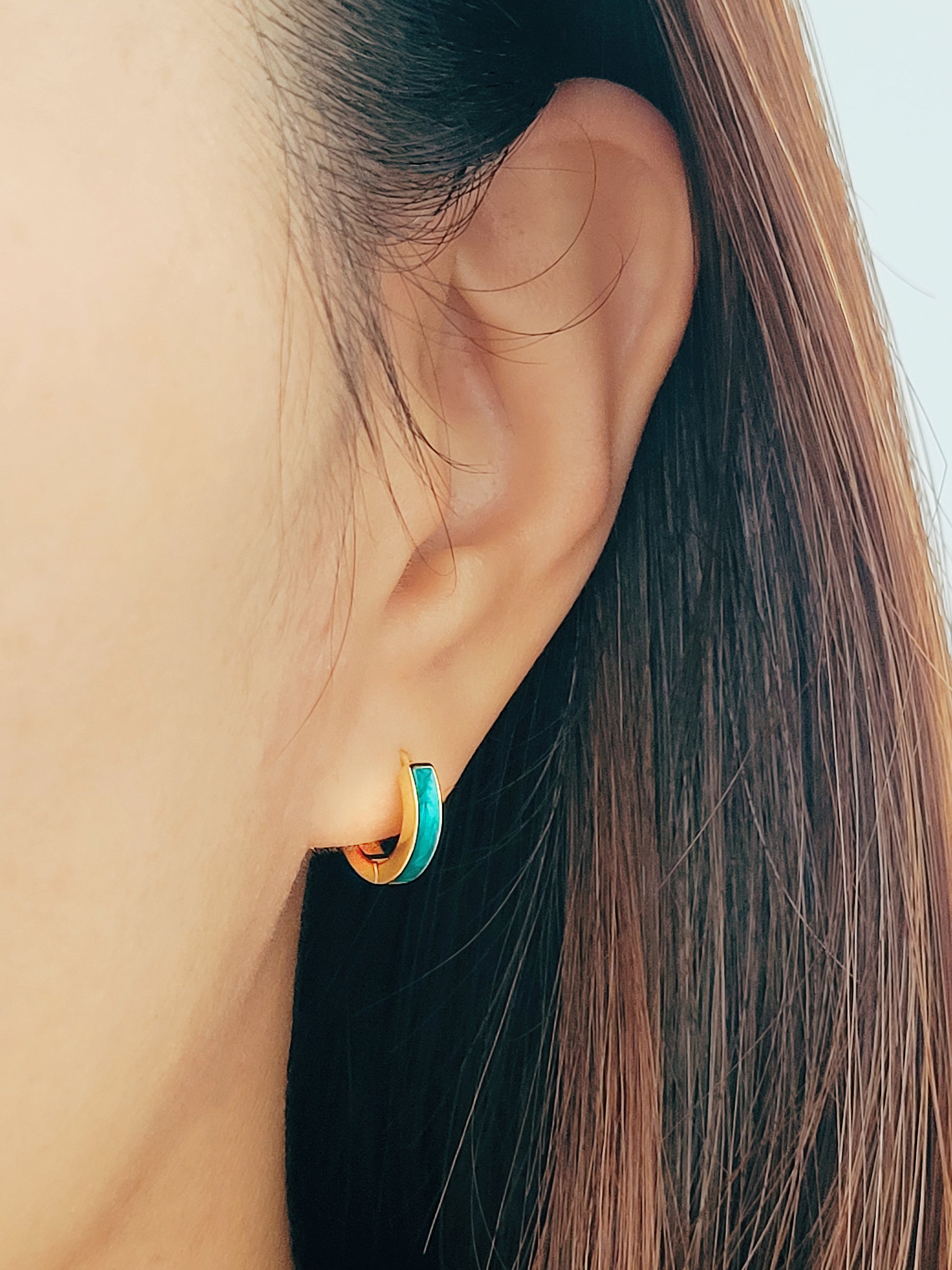 A woman wears eye-catching emeral green enamel huggie hoop earrings, featuring two vibrant colors - turquoise and gold, enhancing her fashionable appearance.