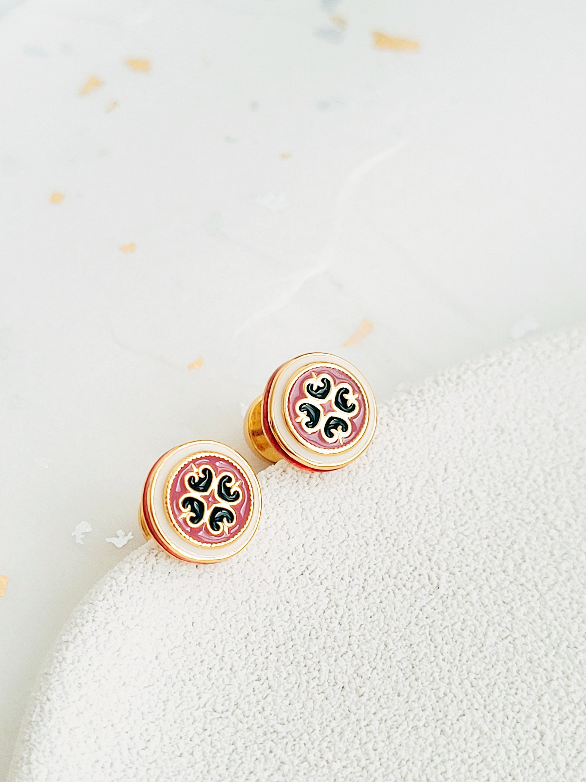 Closeup of retro-style stud earrings featuring a pink and dark green geometric tile pattern with gold accents. These vintage-inspired enamel earrings offer a classic, timeless design adding a touch of elegance to any outfit.