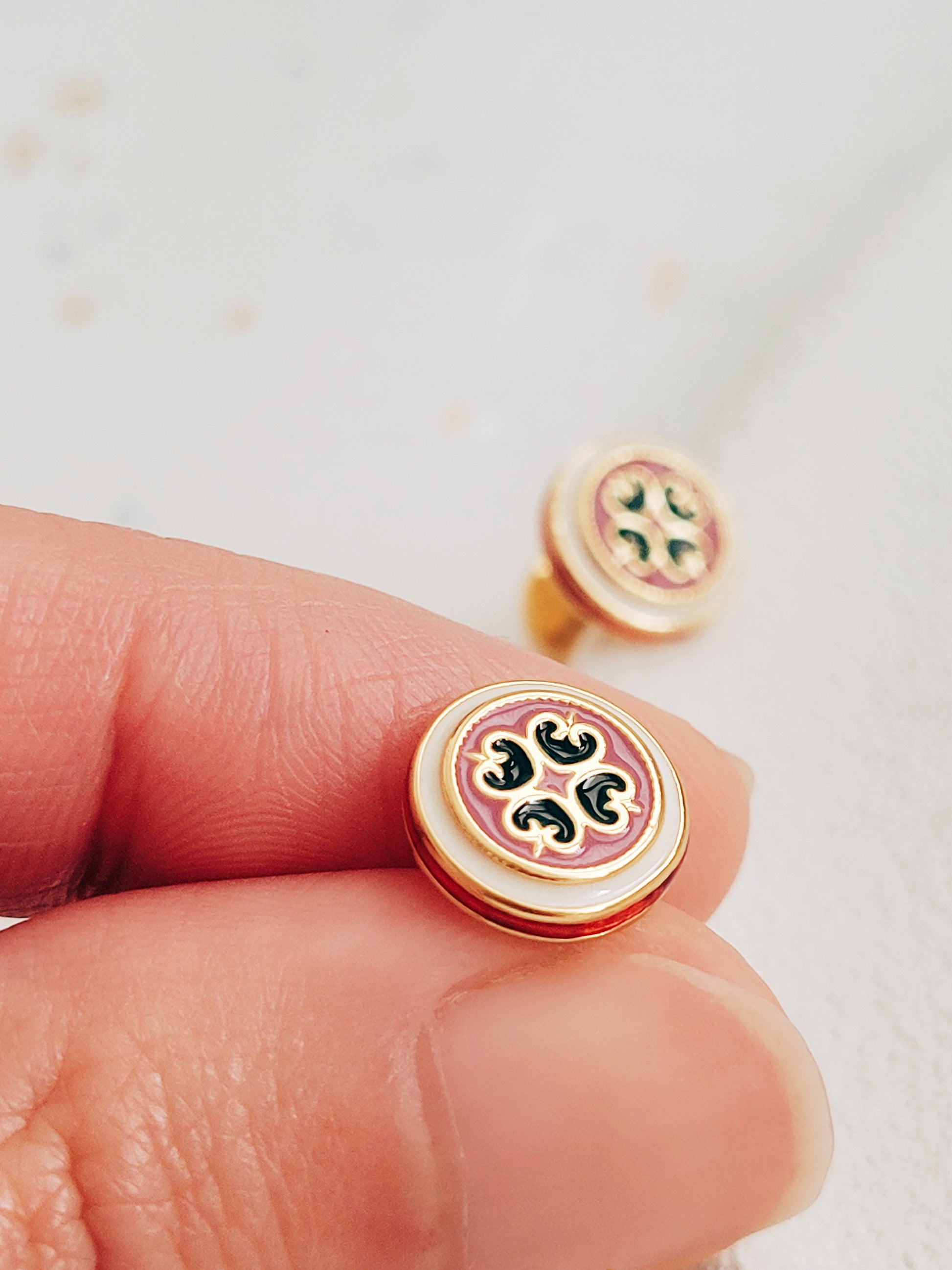 Closeup of retro-style stud earrings featuring a pink and dark green geometric tile pattern with gold accents. These vintage-inspired enamel earrings offer a classic, timeless design adding a touch of elegance to any outfit.