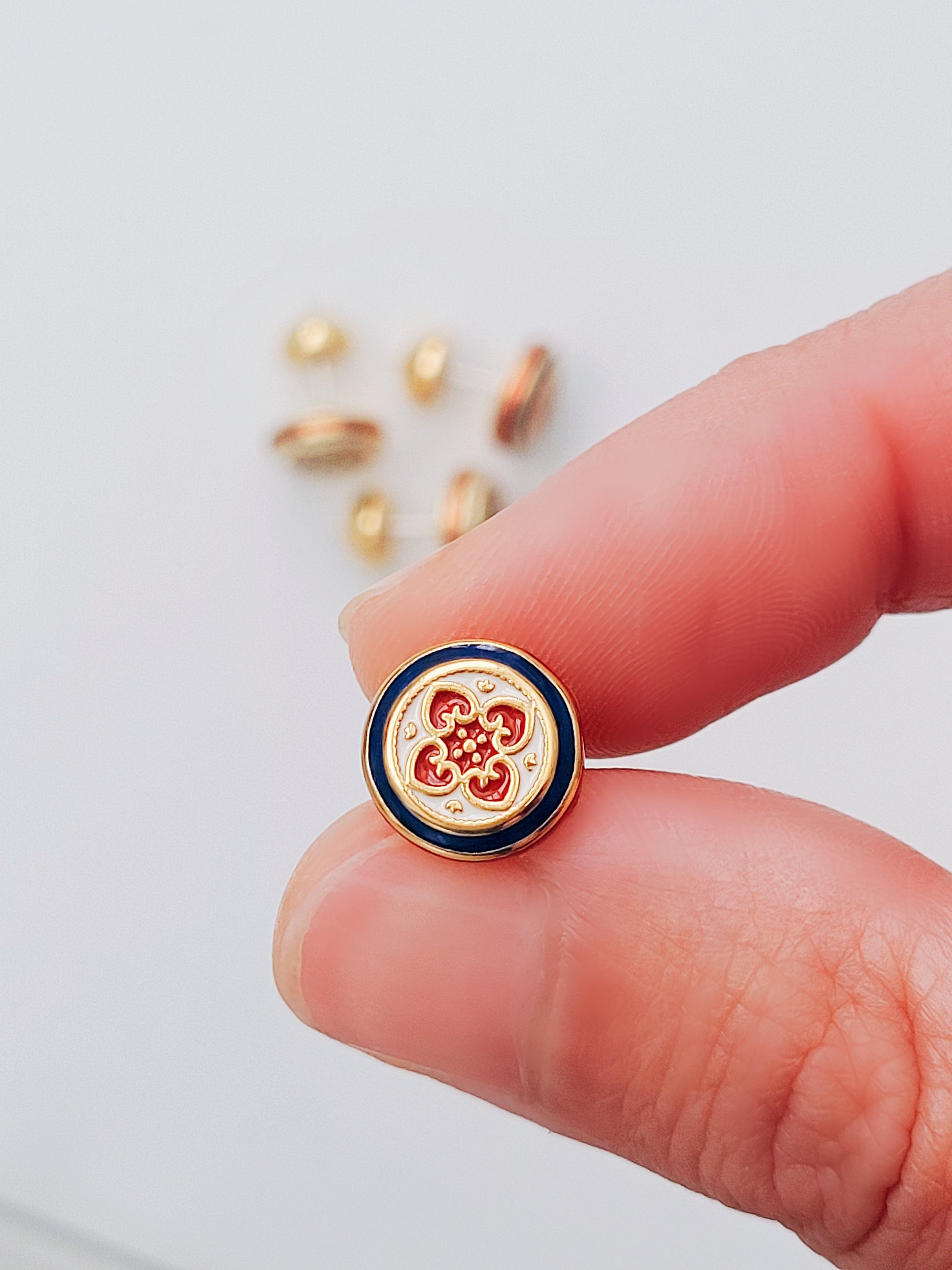 A woman's hand holding a Retro Gothic Enamel Earrings featuring a red and blue vintage inspired design with gold accents on a round ear stud.