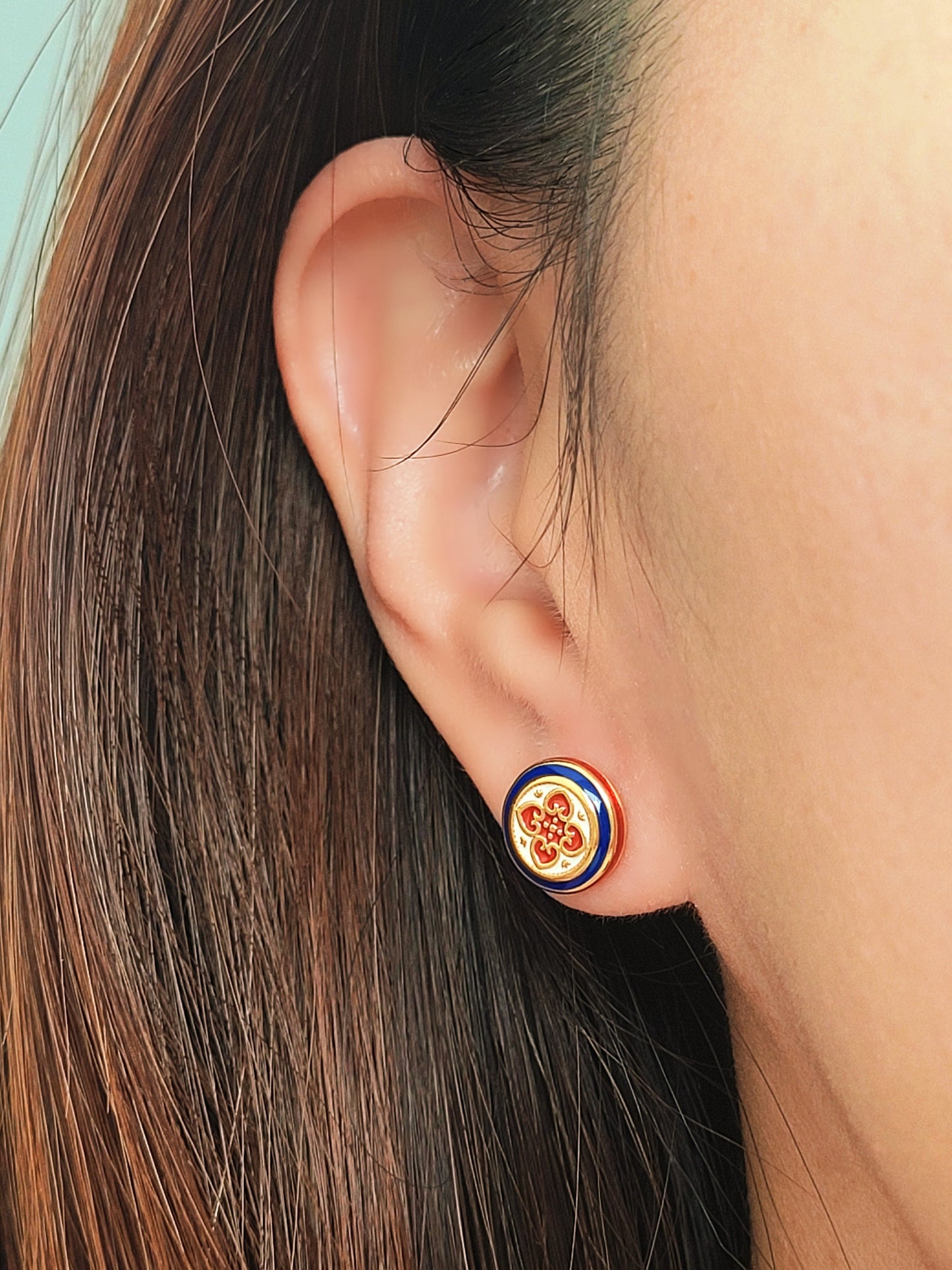 Close-up of a woman's ear wearing a pair of Retro Gothic tile pattern enamel stud earrings in red, blue, and gold. The intricate design features a vintage-style pattern with a bold color combination, creating a striking and elegant look."