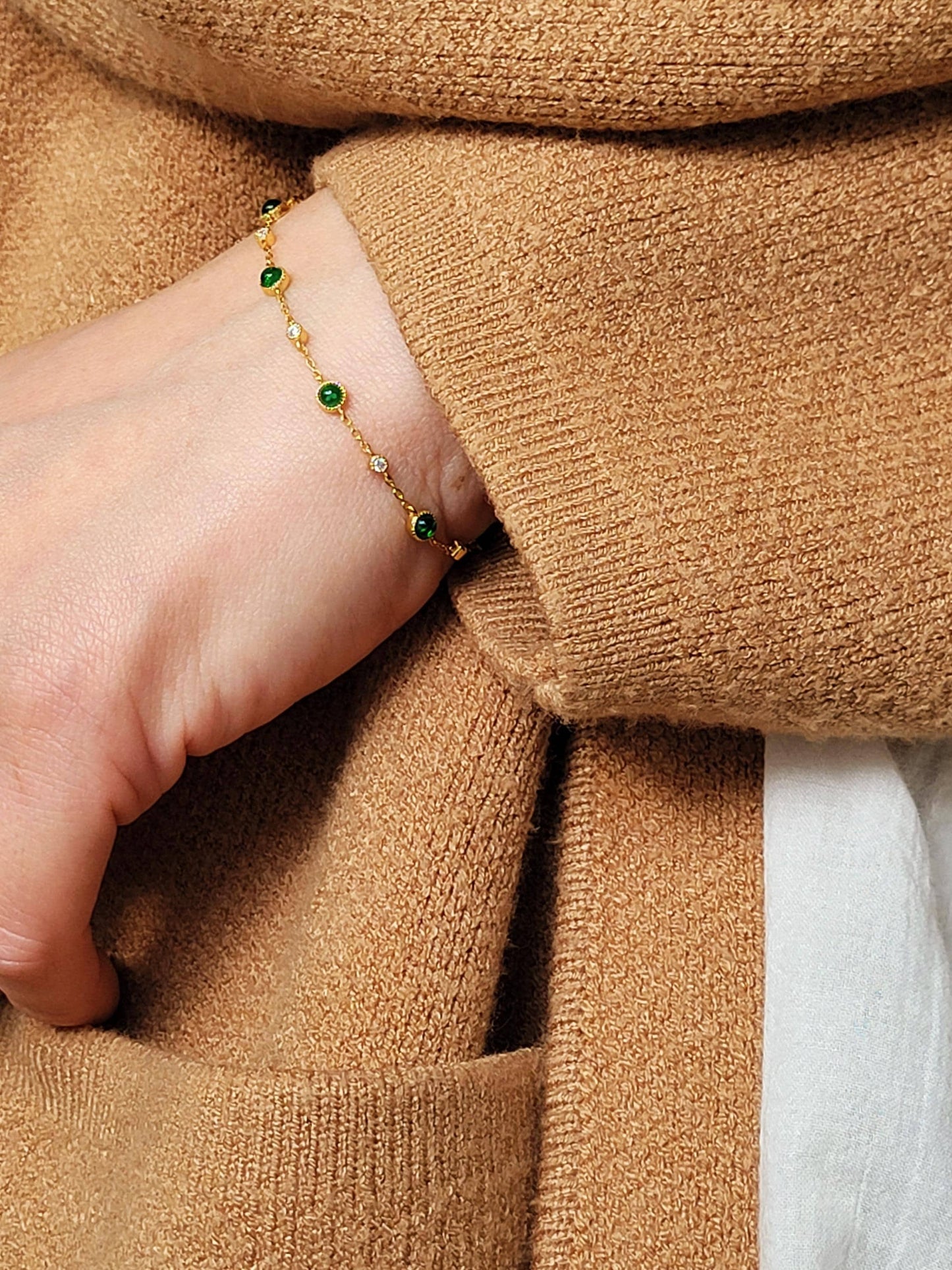 Close-up of a hand wearing a minimalist gold bracelet with green beads, against a brown fabric background.