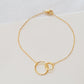 A delicate gold chain bracelet with two interlocking textured circular rings at the center, displayed on a light off-white textured fabric background.