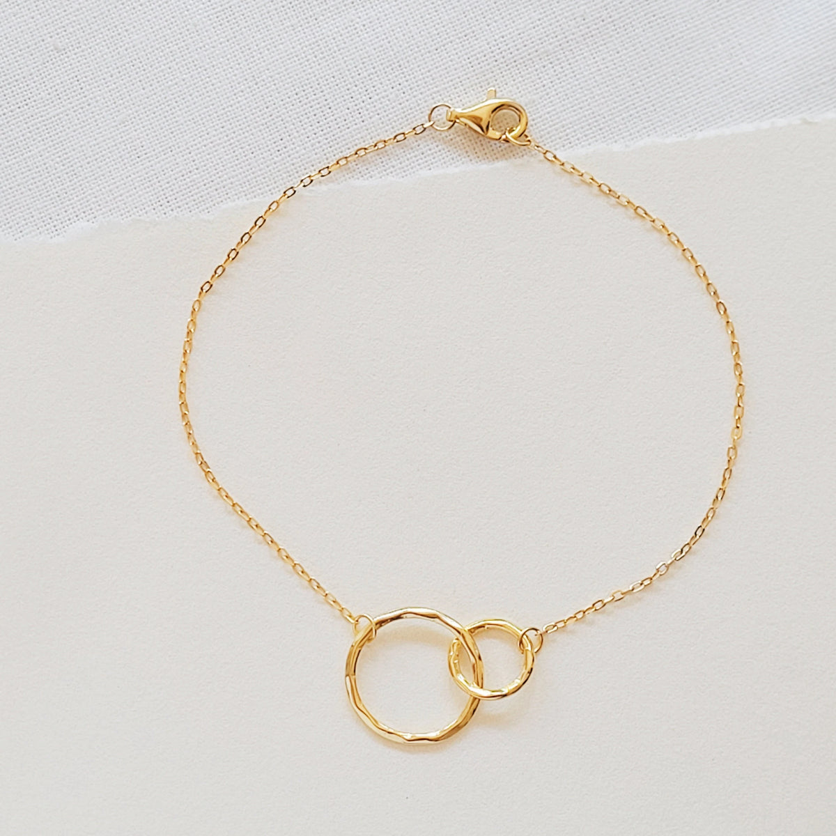 A delicate gold chain bracelet with two interlocking textured circular rings at the center, displayed on a light off-white textured fabric background.