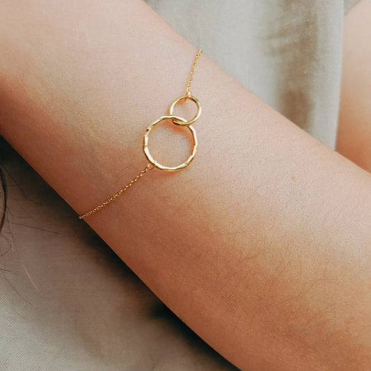A delicate gold chain bracelet with two interlocking textured rings worn on a person's wrist, shown against a soft neutral background.