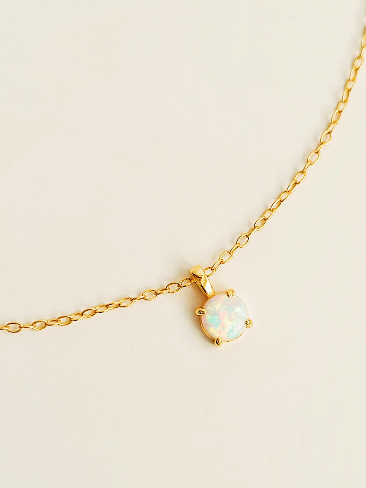 A delicate gold chain necklace featuring a small oval opal pendant in a gold bezel setting, displayed on a clean, light beige background.