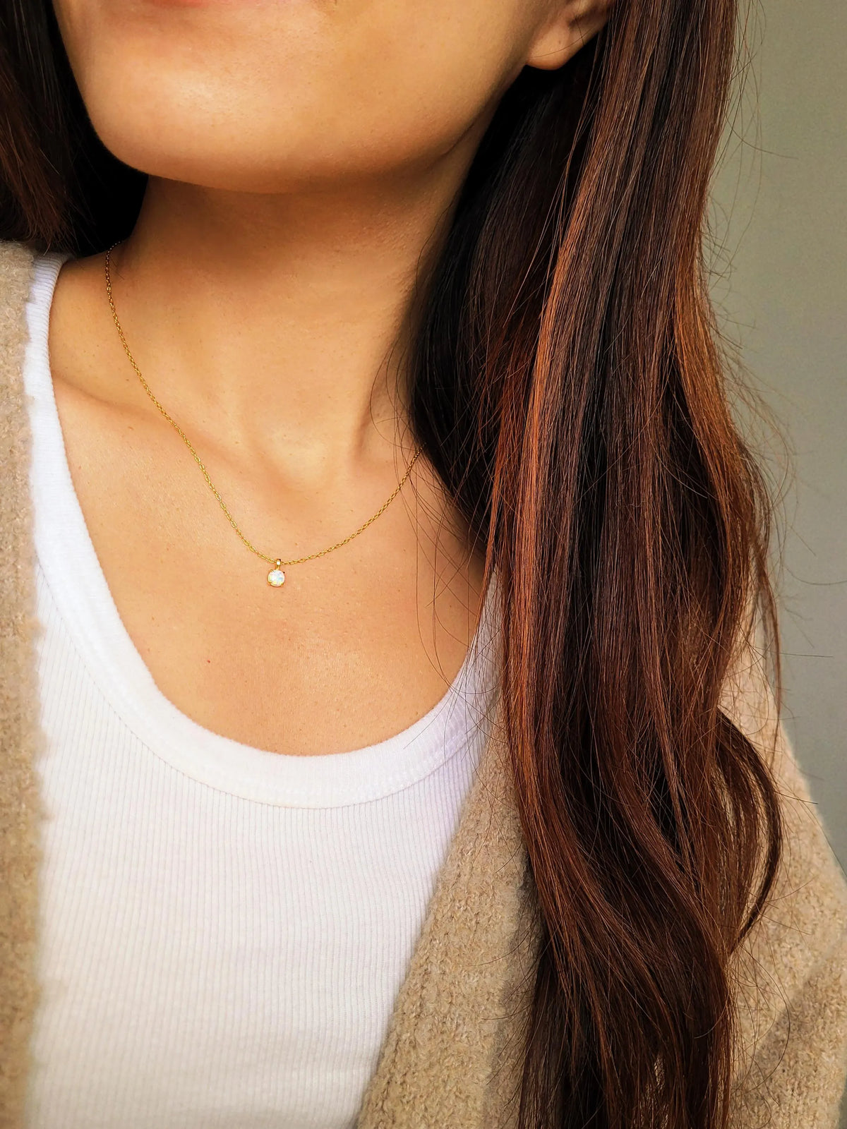 A lifestyle shot of a woman wearing a delicate gold chain necklace with a small white Opal pendant, styled over a white ribbed tank top and a beige cardigan.