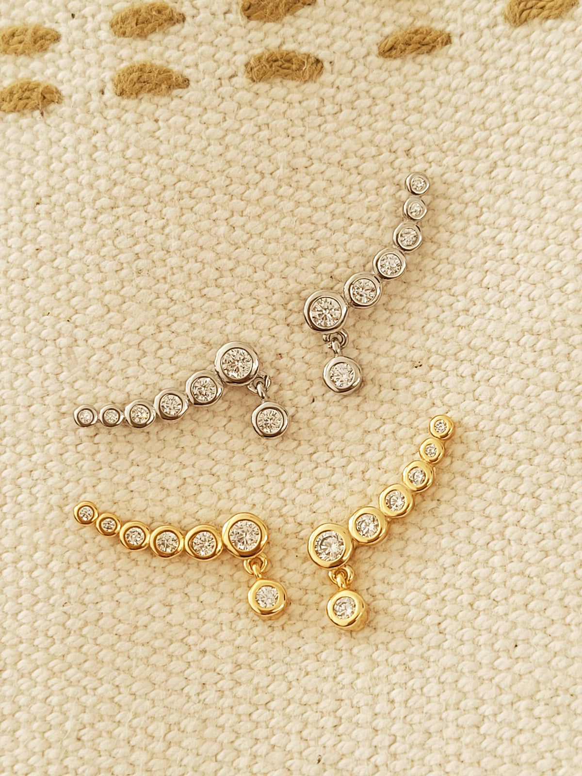 Gold and silver bubble climber earrings displayed together on textured beige fabric, showing the two metal finishes.