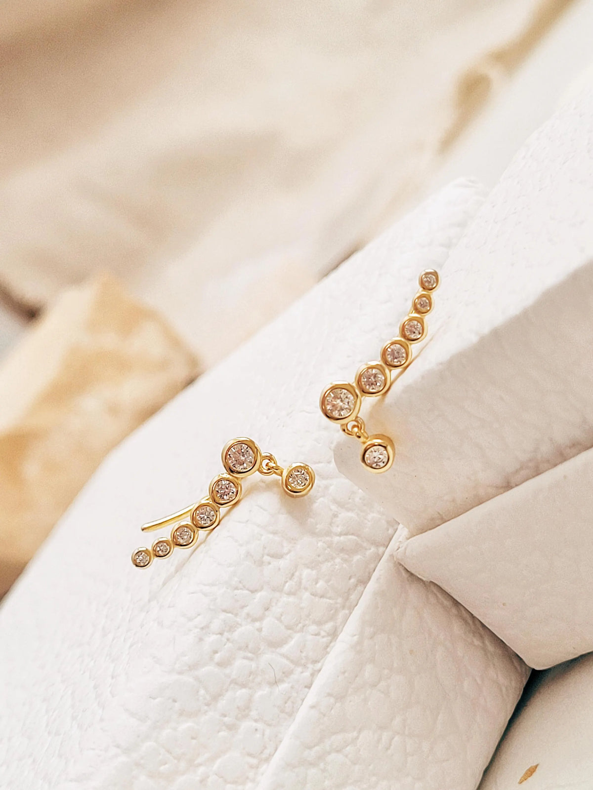 Pair of gold-plated bubble climber earrings with clear cubic zirconia stones arranged on soft white textured jewellery box.