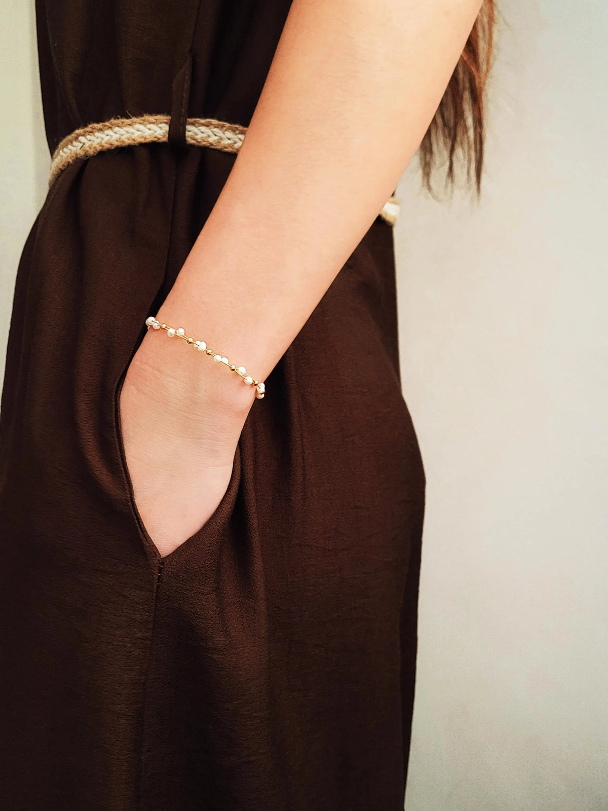 A close-up of a woman wearing a delicate pearl bracelet on her wrist, paired with a dark brown dress and a woven belt.
