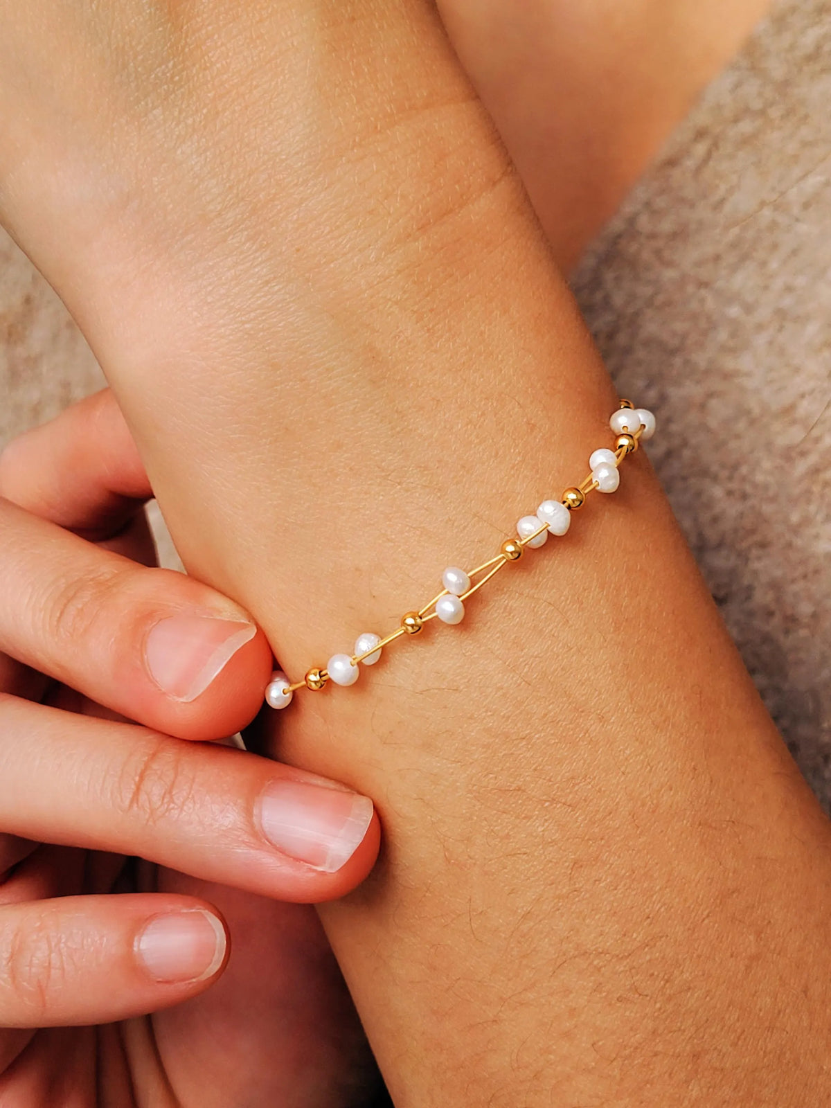 A delicate gold chain bracelet adorned with small tiny pearls, shown worn on a woman's wrist with fingers gently touching the band.