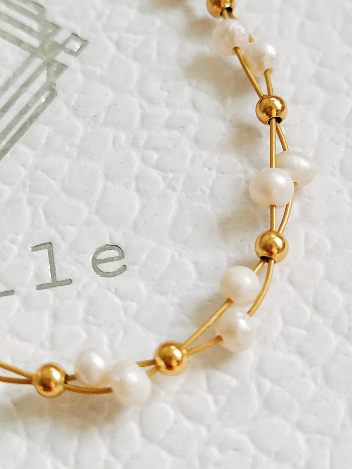 A close-up of a delicate gold wire bracelet with alternating white pearl beads and small gold spacer beads, laid on a textured white surface.