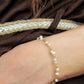 A close-up of a woman's wrist wearing a delicate bracelet made of white pearls and small gold beads, set against a dark brown textured garment with a braided beige rope detail.