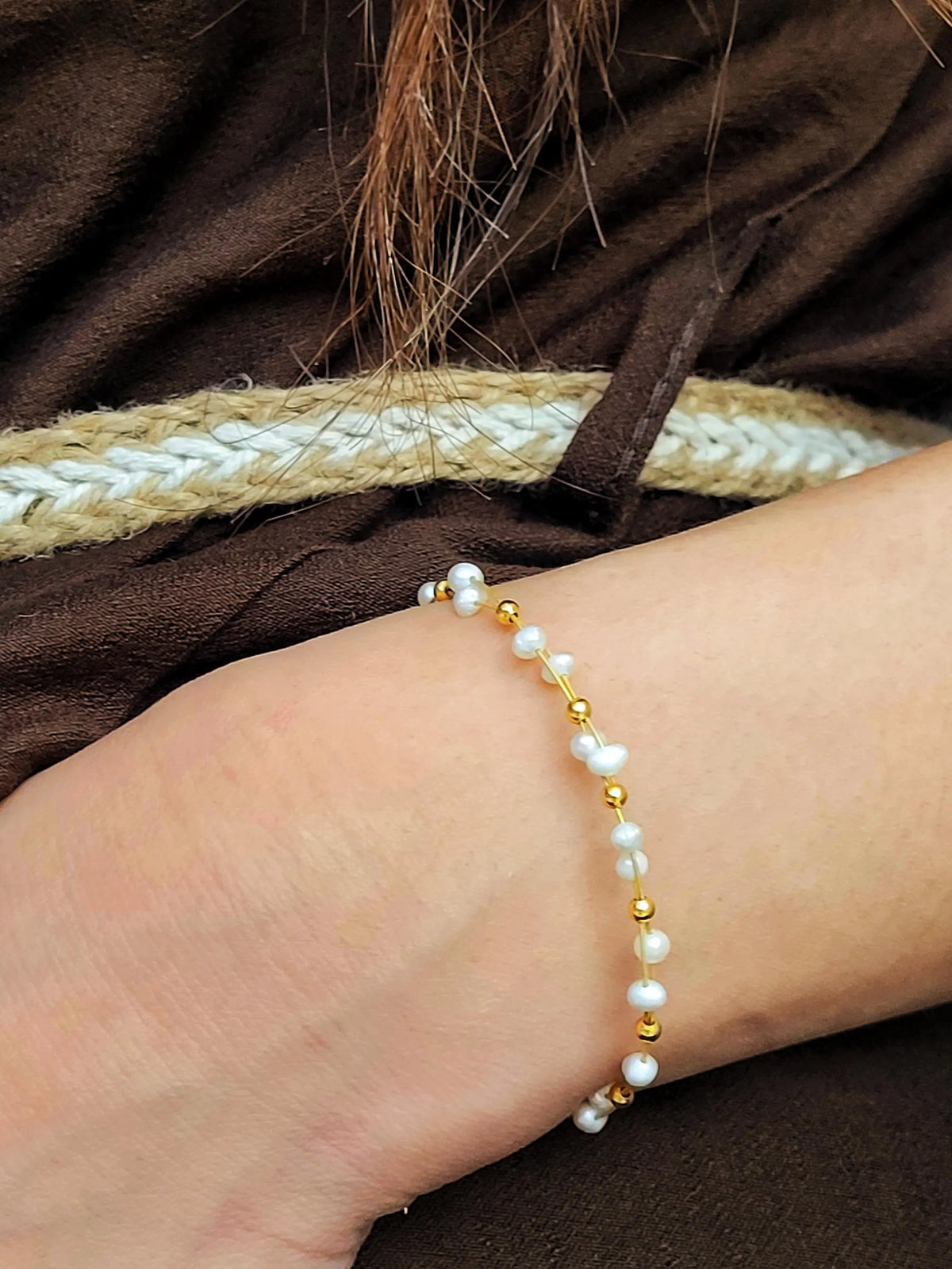 A close-up of a woman's wrist wearing a delicate bracelet made of white pearls and small gold beads, set against a dark brown textured garment with a braided beige rope detail.