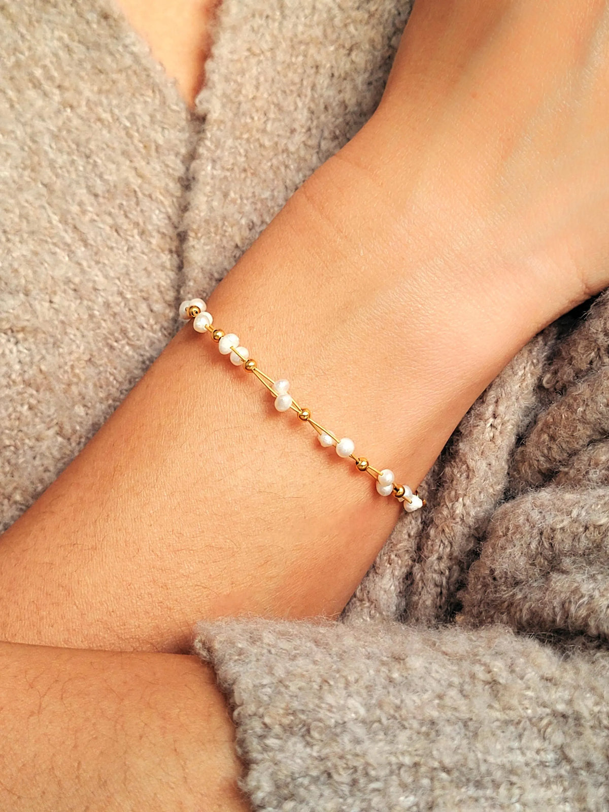 A delicate gold chain bracelet with small white pearl beads, shown worn on a person's wrist against a cozy gray knit sweater.