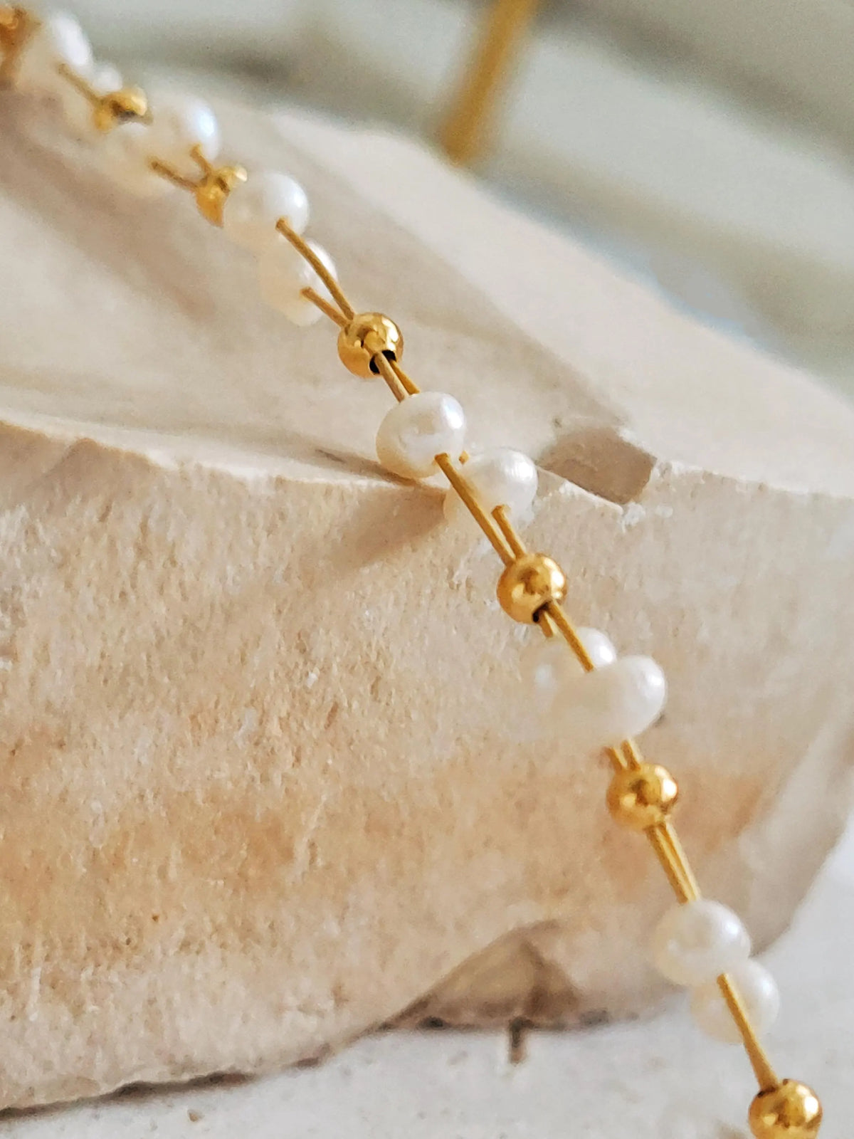 A close-up of a beaded bracelet with white freshwater pearls round beads and small polished gold spacer beads, draped over a light stone block.