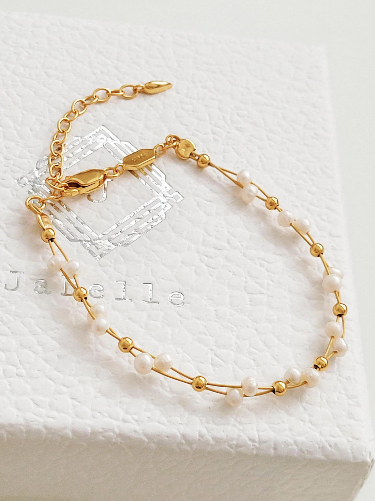 A delicate gold chain bracelet with small white freshwater pearls, displayed on a textured white JaBelle jewelry box. the box.
