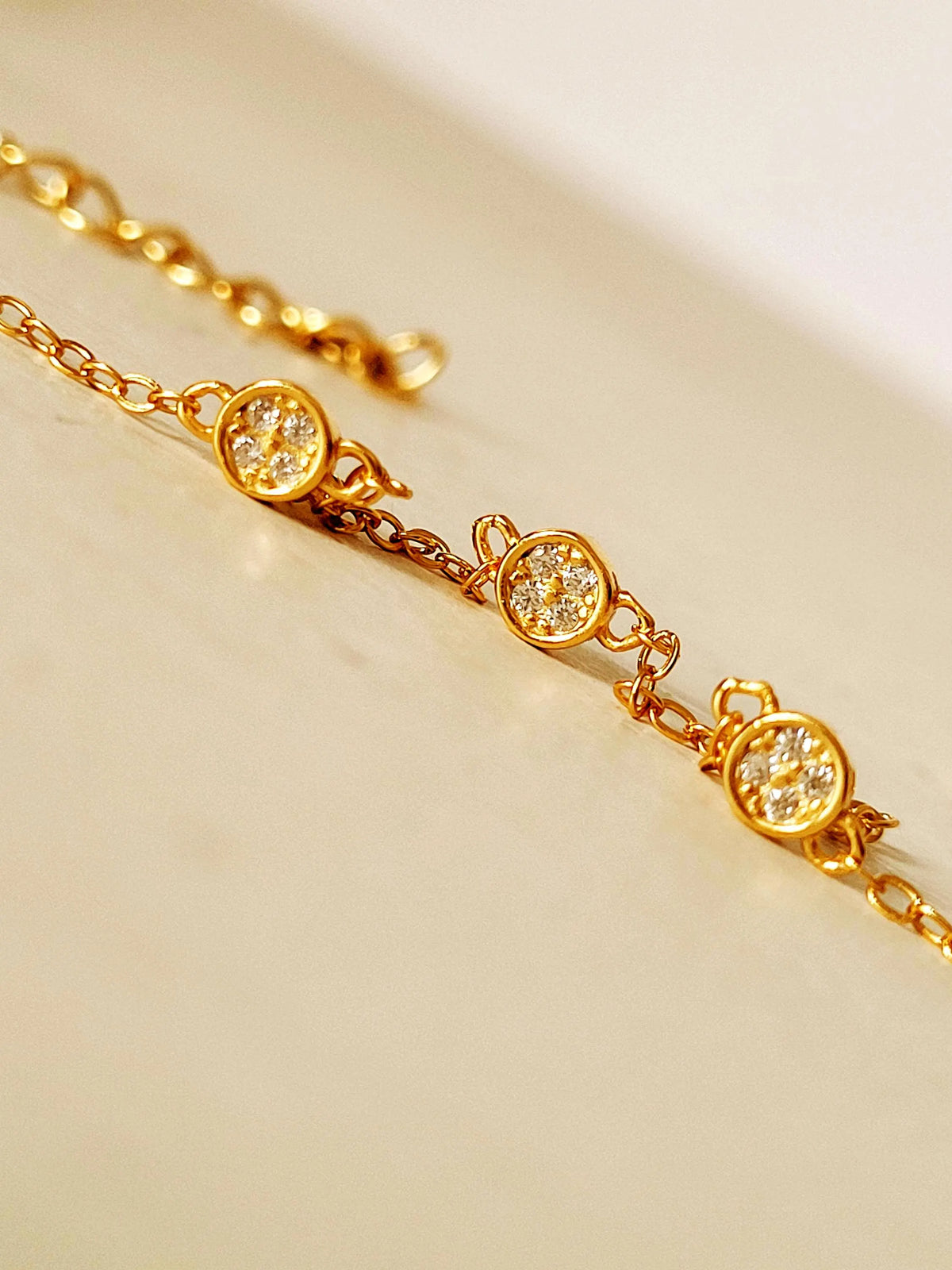 A gold chain bracelet with three oval bezel-set clear stones, photographed close-up on a warm cream surface.