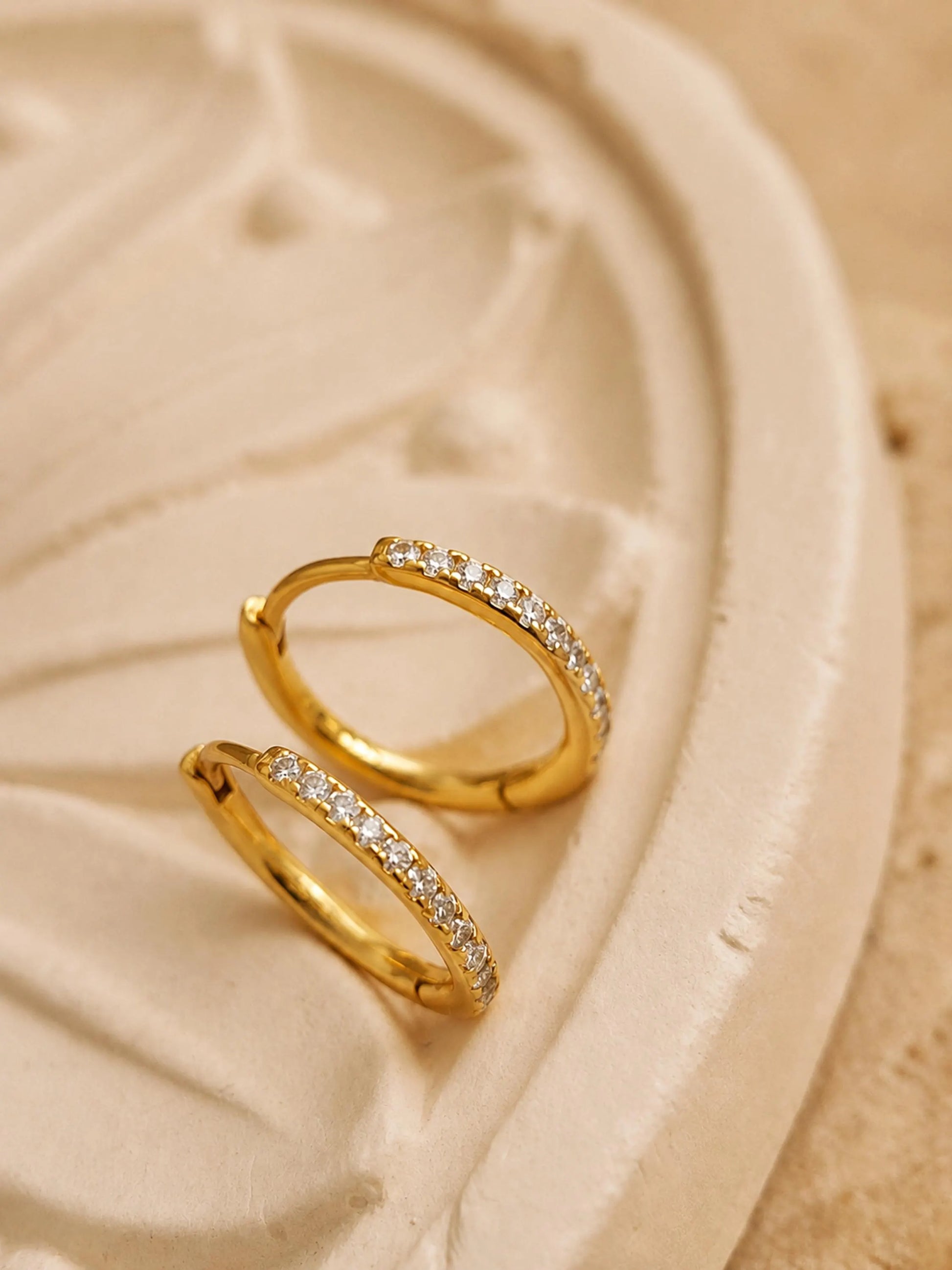 A pair of thin, hinged huggie hoop earrings in gold vermeil featuring a row of sparkling, clear cubic zirconia stones displayed on a luxury beige flower plate.