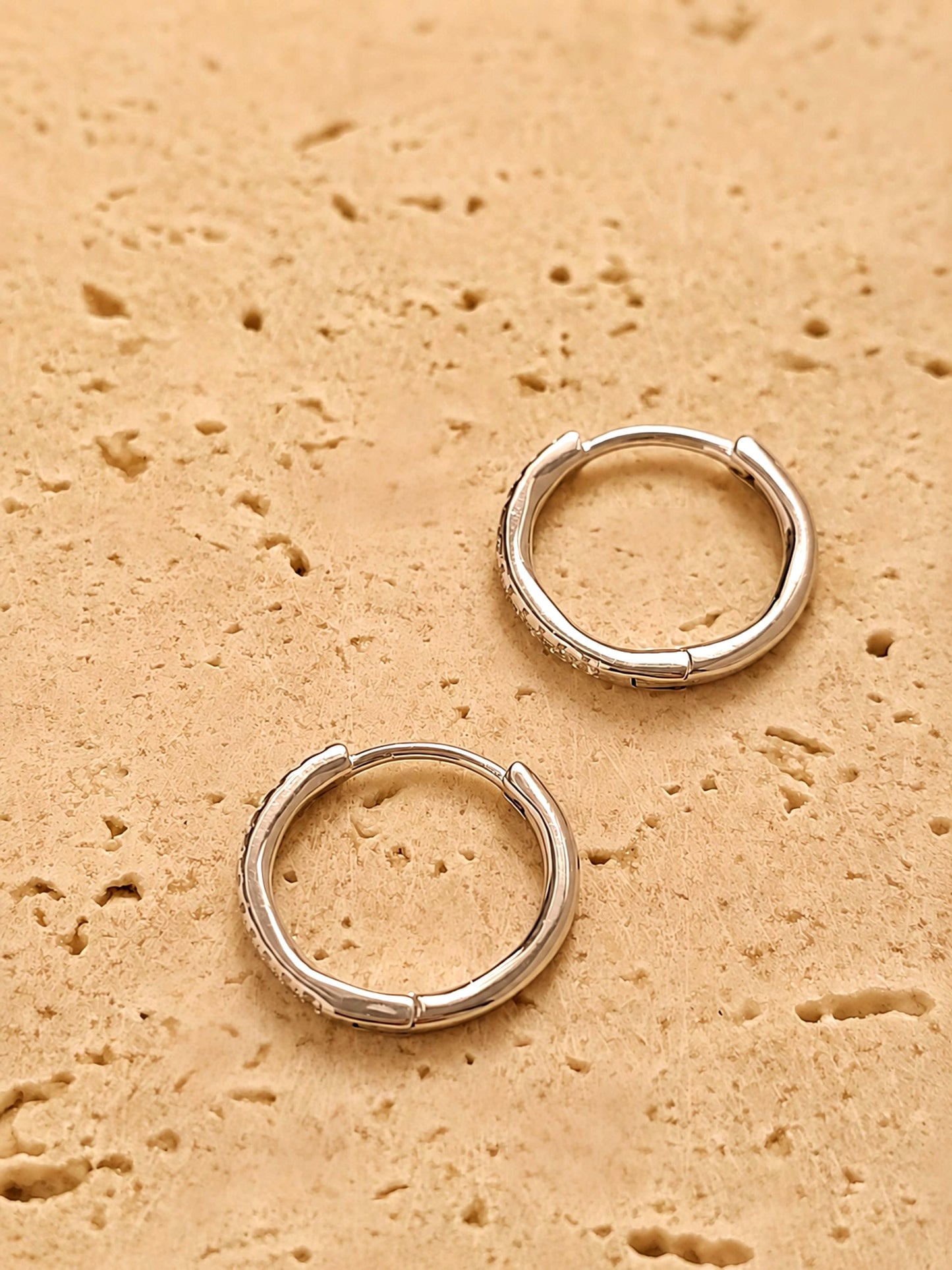 A pair of dainty gold huggie earrings featuring irregular organic shape with hinged snap closure on a beige color stone surface.