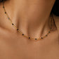 A delicate gold necklace with small, dark beads spaced along a fine chain. The necklace is shown against a neutral skin tone, highlighting its simple elegance. The chain has a twisted rope design.