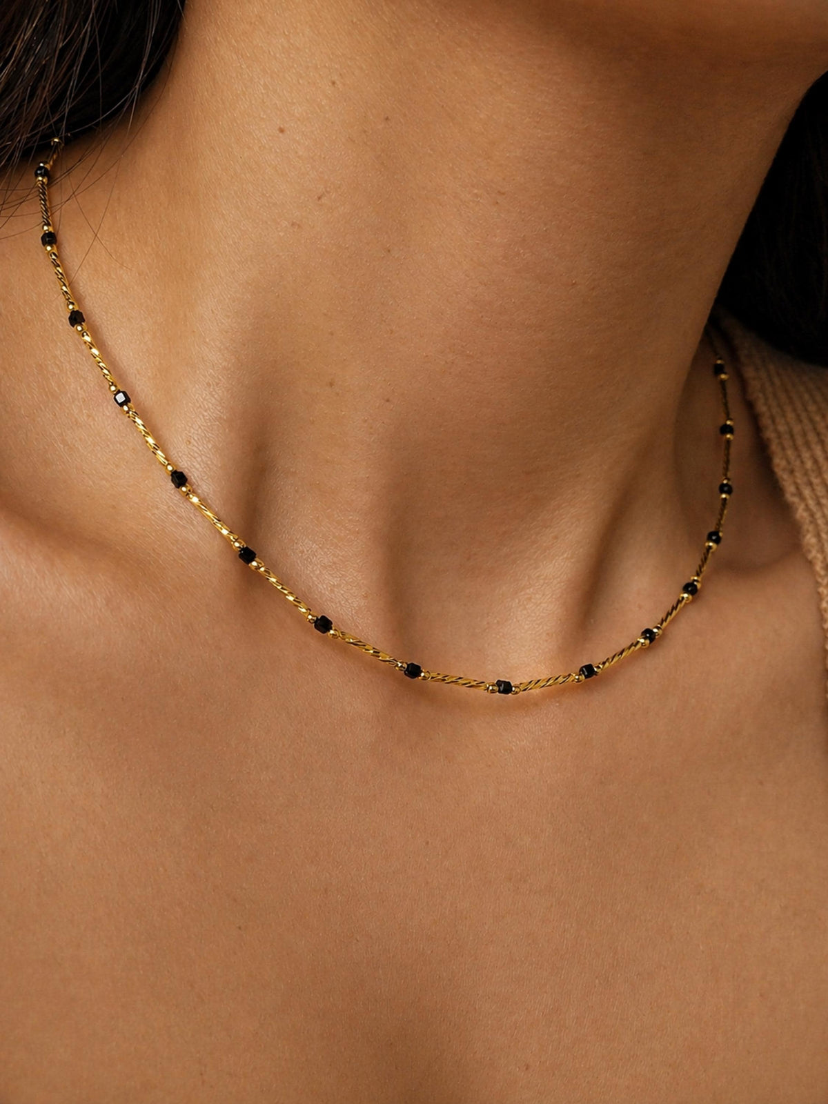 A delicate gold necklace with small, dark beads spaced along a fine chain. The necklace is shown against a neutral skin tone, highlighting its simple elegance. The chain has a twisted rope design.