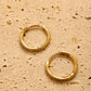 A pair of dainty gold huggie earrings featuring irregular organic shape with hinged snap closure on a beige color textured stone surface.