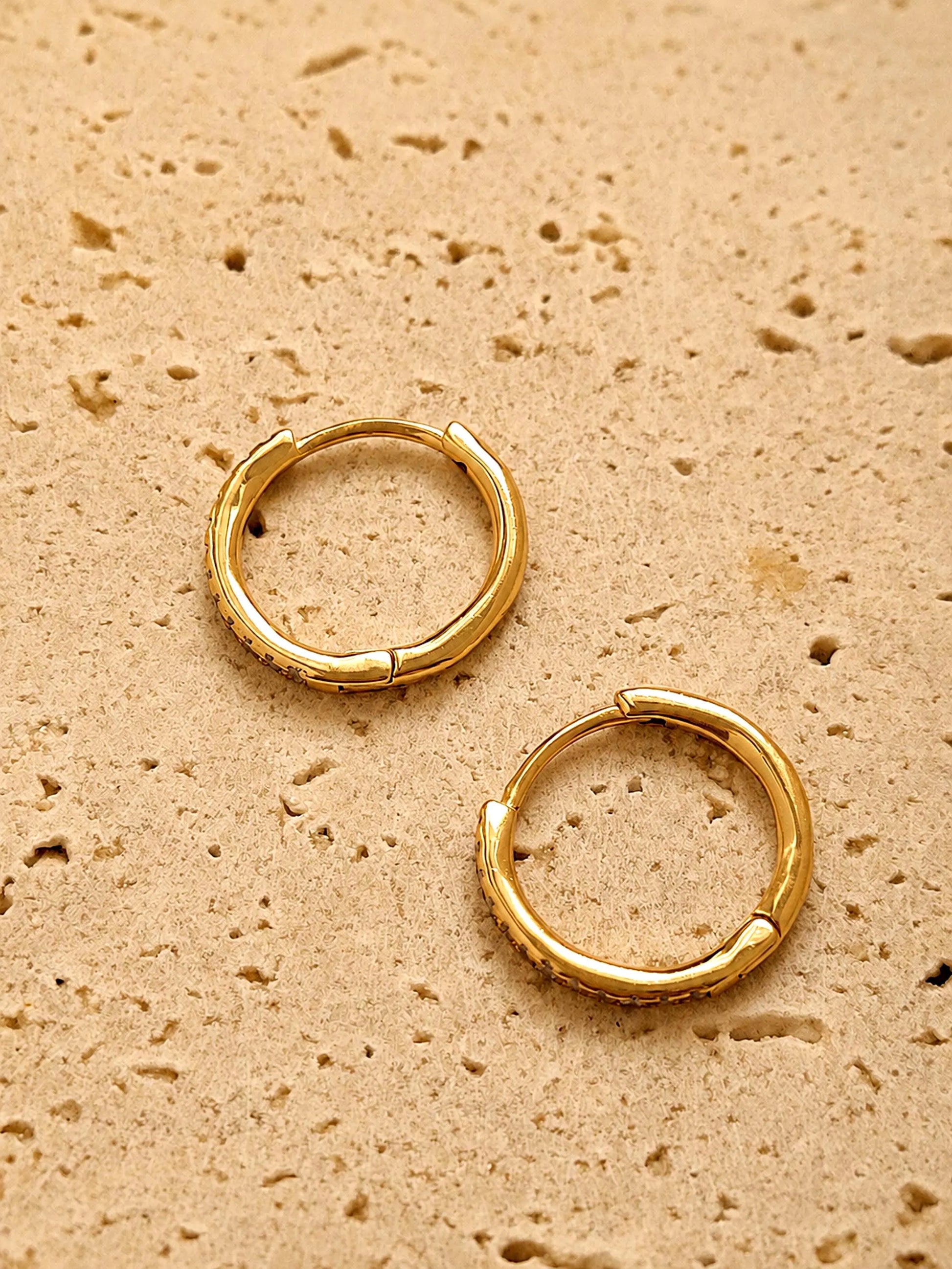 A pair of dainty gold huggie earrings featuring irregular organic shape with hinged snap closure on a beige color textured stone surface.