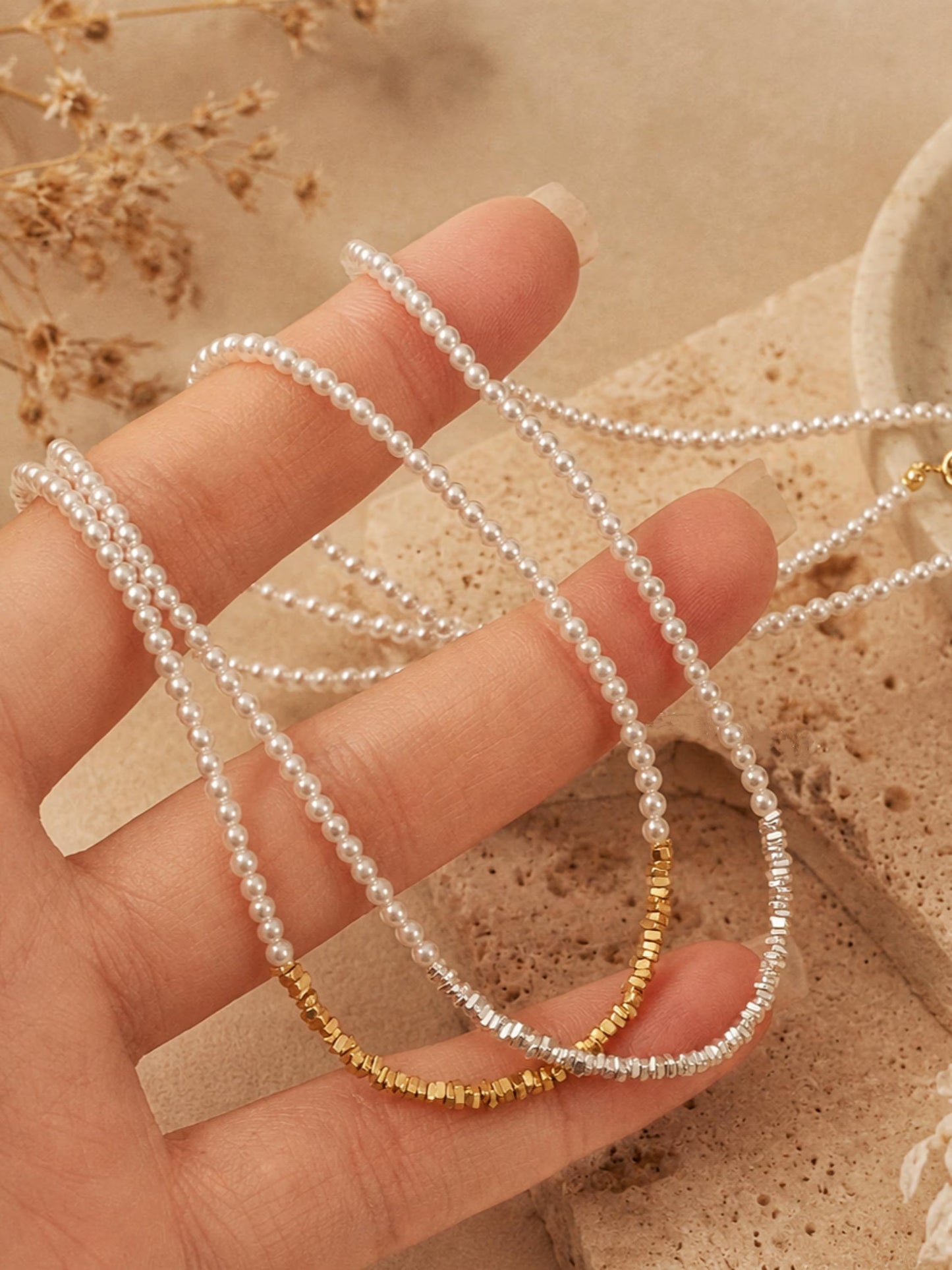 Hand holding a beaded choker necklace with gold and silver beads against a beige background.